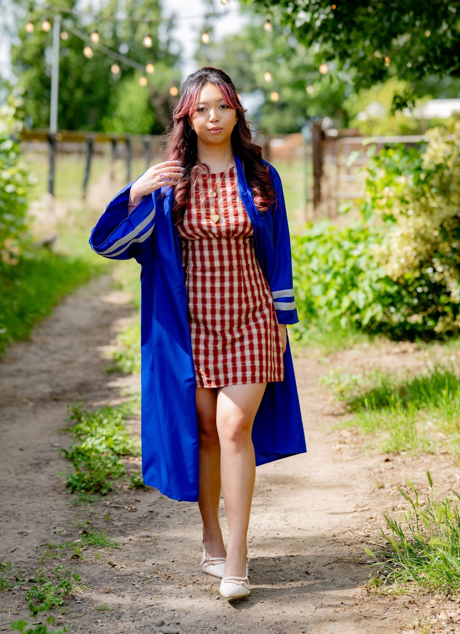 Young woman in blue graduation gown over red gingham dress strolls along a lush green garden path.