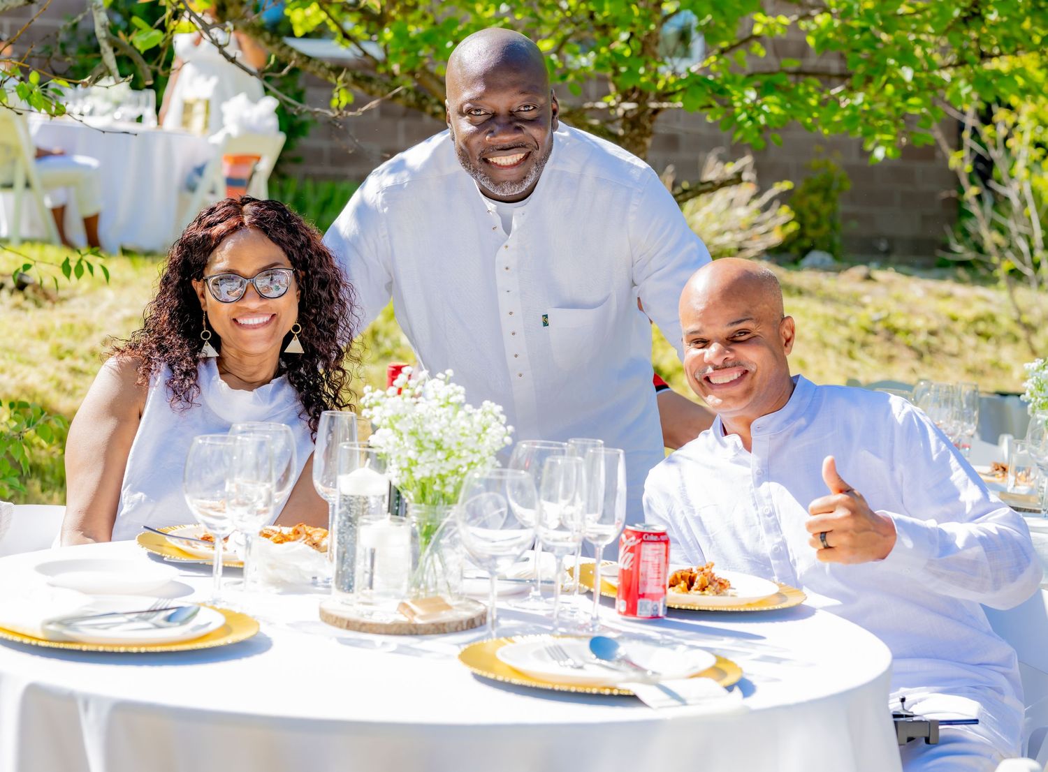 Three people smiling at a elegantly set outdoor dining table with white linens and flowers.