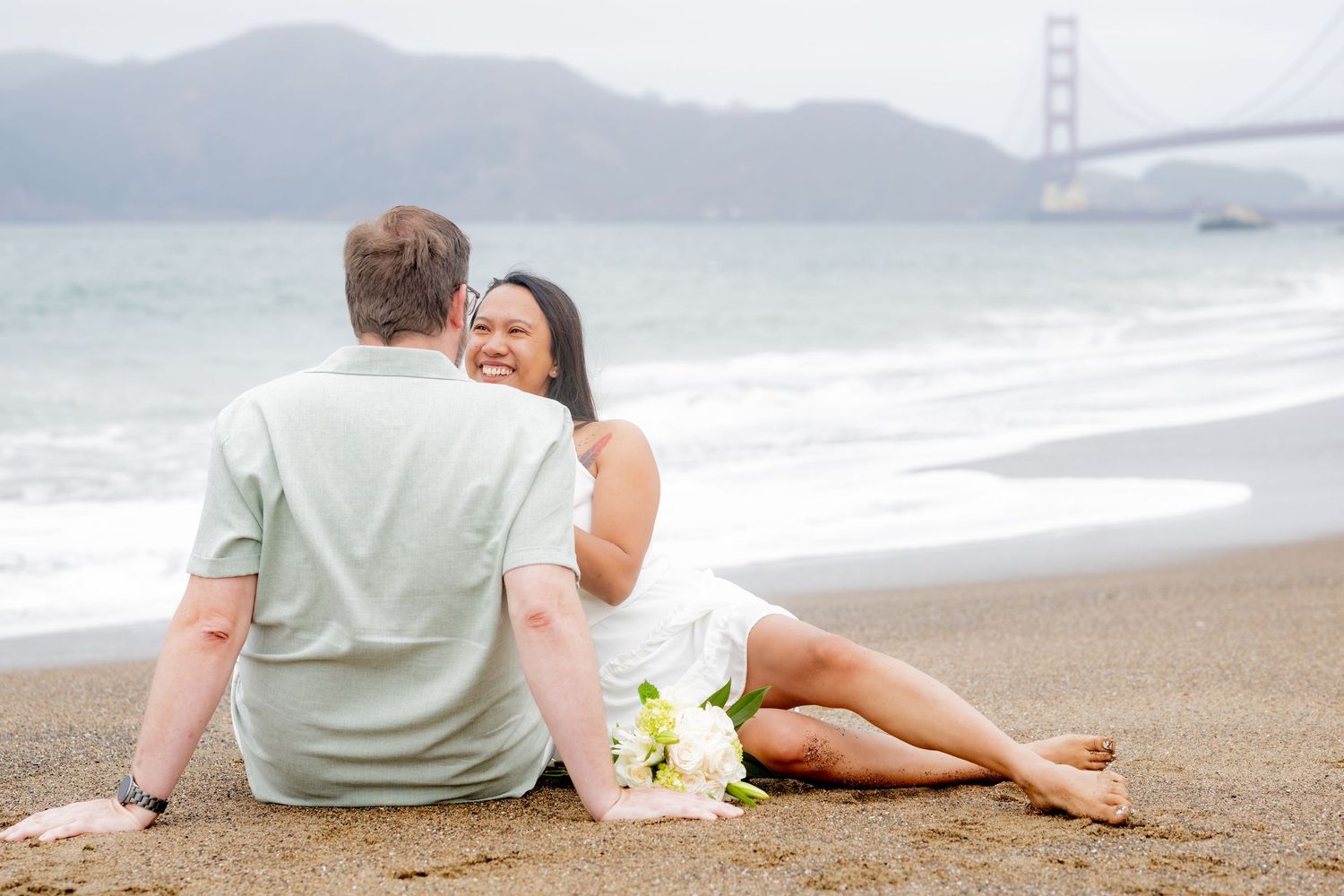 Couple sitting together on Baker Beach sand gazing at the Golden Gate Bridge with a bouquet nearby.