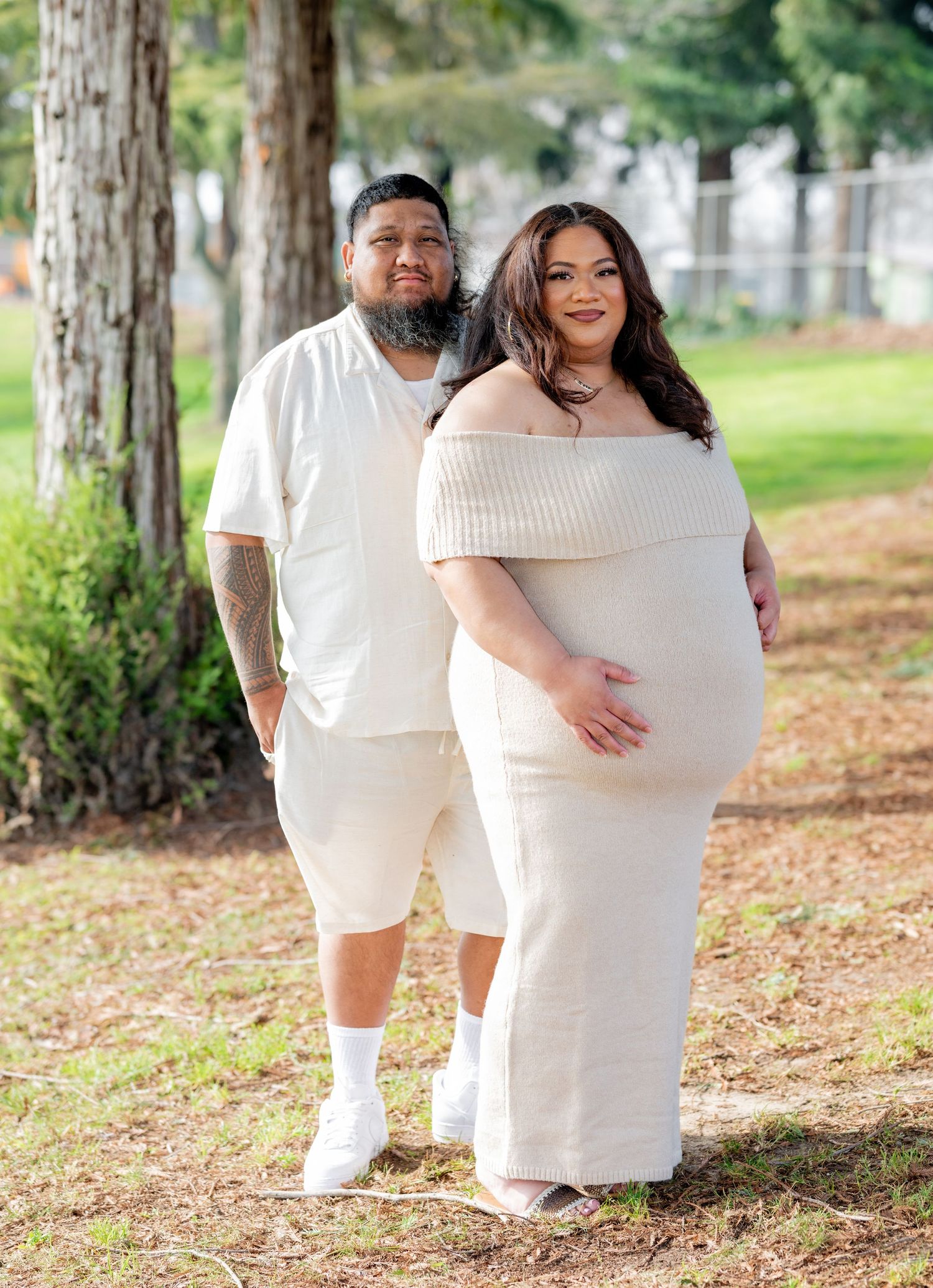 Couple in coordinating cream outfits pose together in a park, woman in off-shoulder maternity dress showing baby bump.
