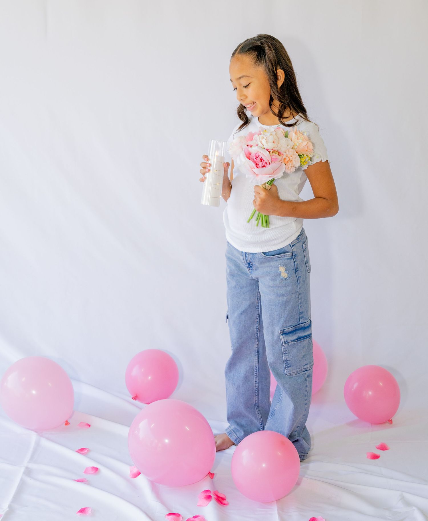 Young girl holds pink flowers and a bottle surrounded by pink balloons on white backdrop.