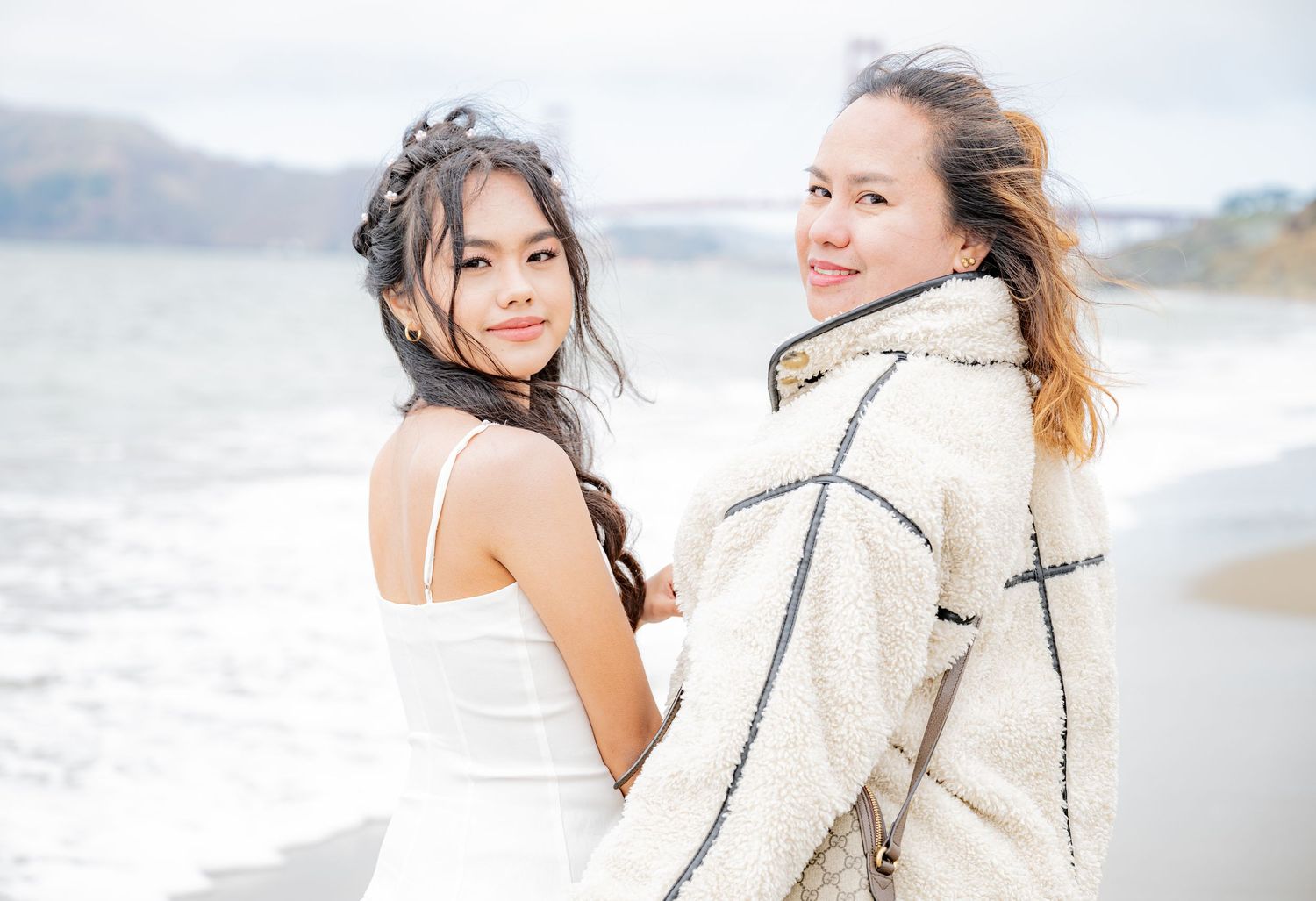 Two smiling young women pose back-to-back on a beach with ocean waves in the background.