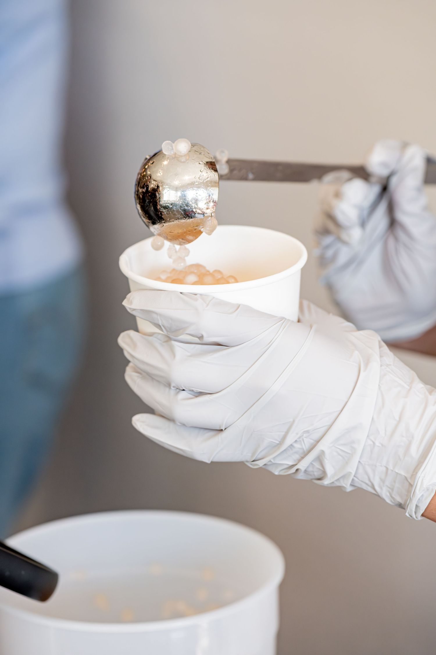 Gloved hands serving soup into a white paper cup with a metal ladle at a food service event.