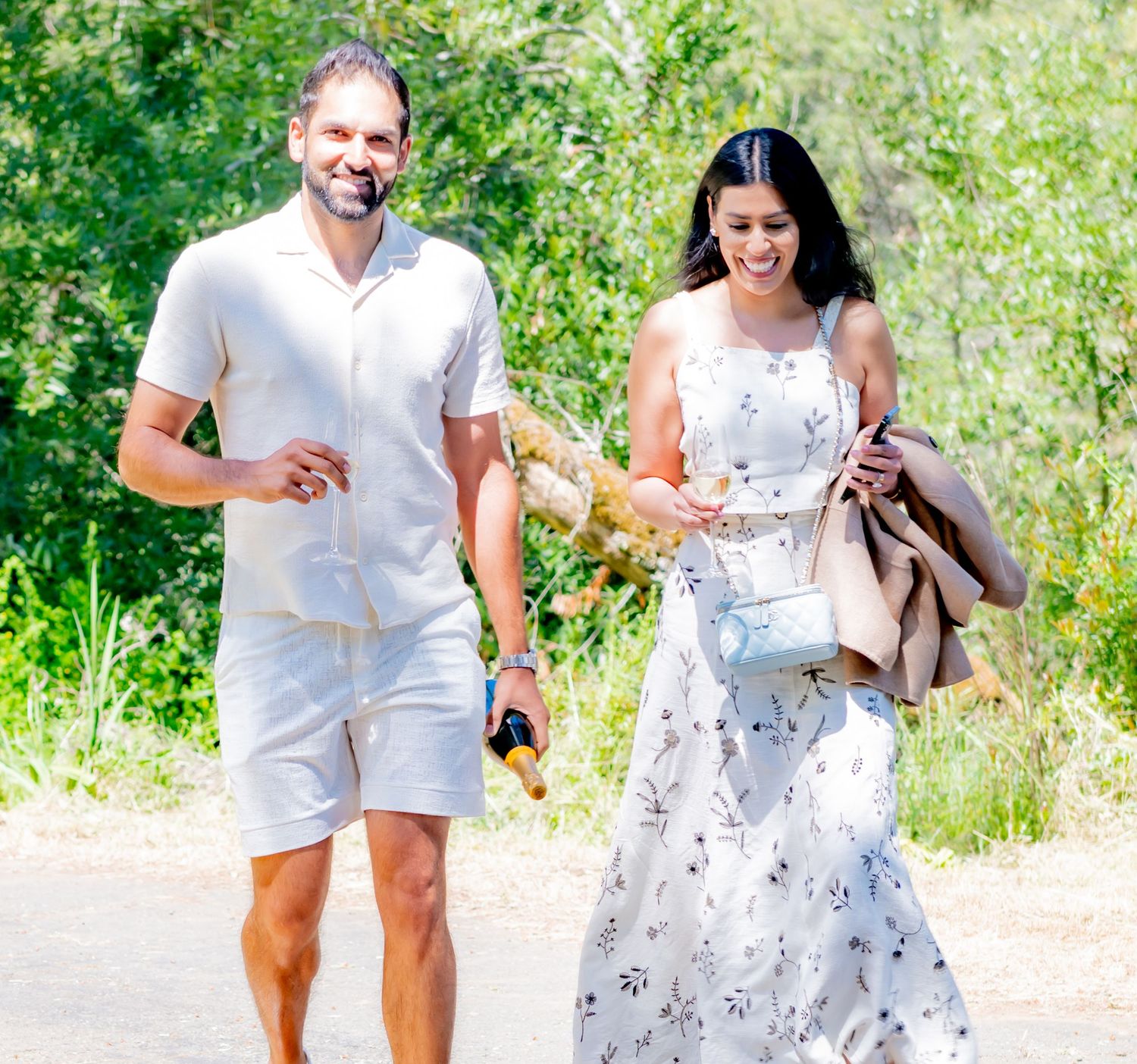 Smiling couple walking outdoors on a sunny path, man in beige outfit, woman in floral dress.