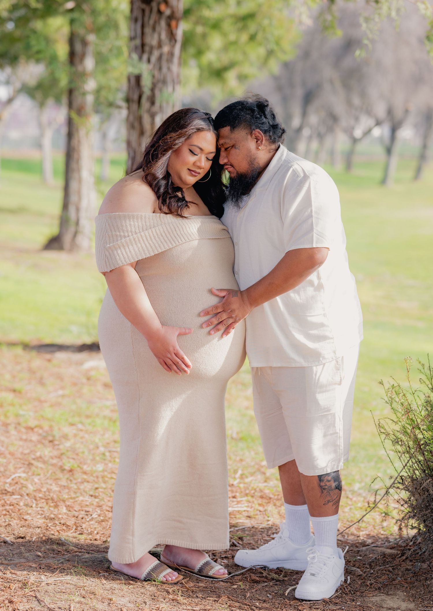 Expecting couple in matching cream outfits tenderly cradling baby bump during outdoor maternity photoshoot.