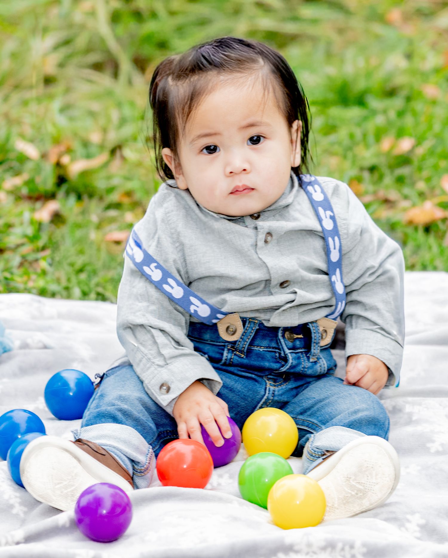 Cute baby boy in suspenders sits on white blanket surrounded by colorful plastic balls outdoors.