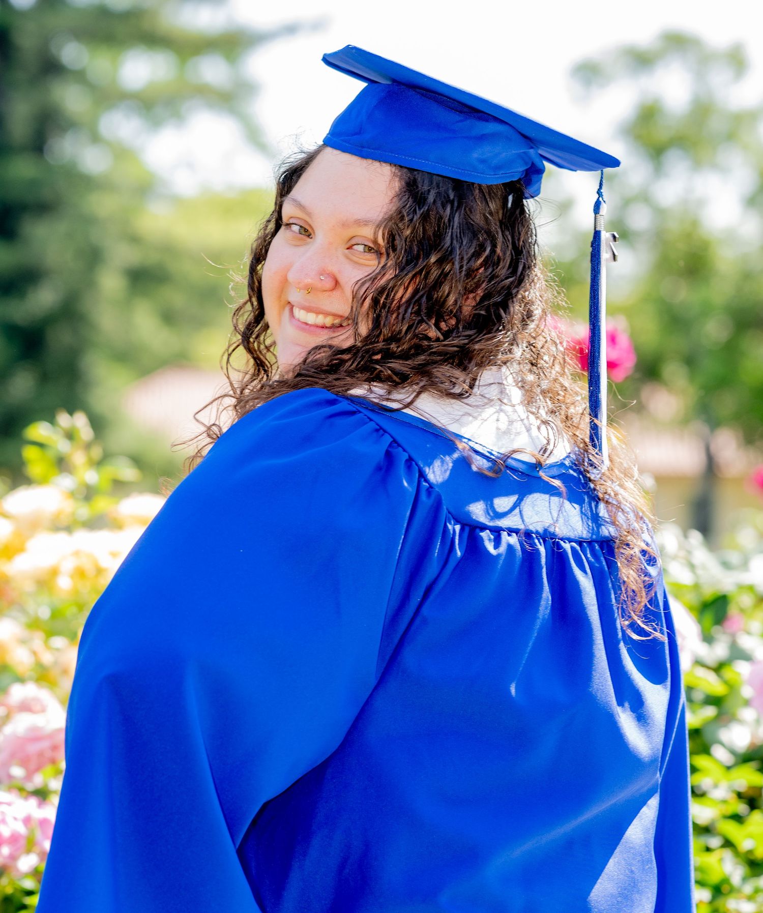 Graduate in blue cap and gown looks over shoulder smiling among colorful garden flowers.