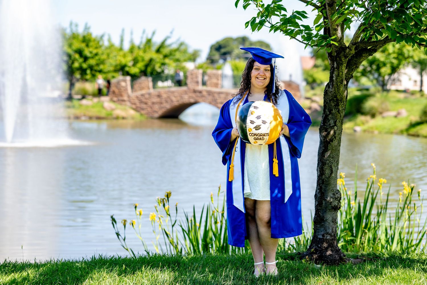 Graduate in blue regalia holds balloon bouquet beside a tranquil pond with fountain.