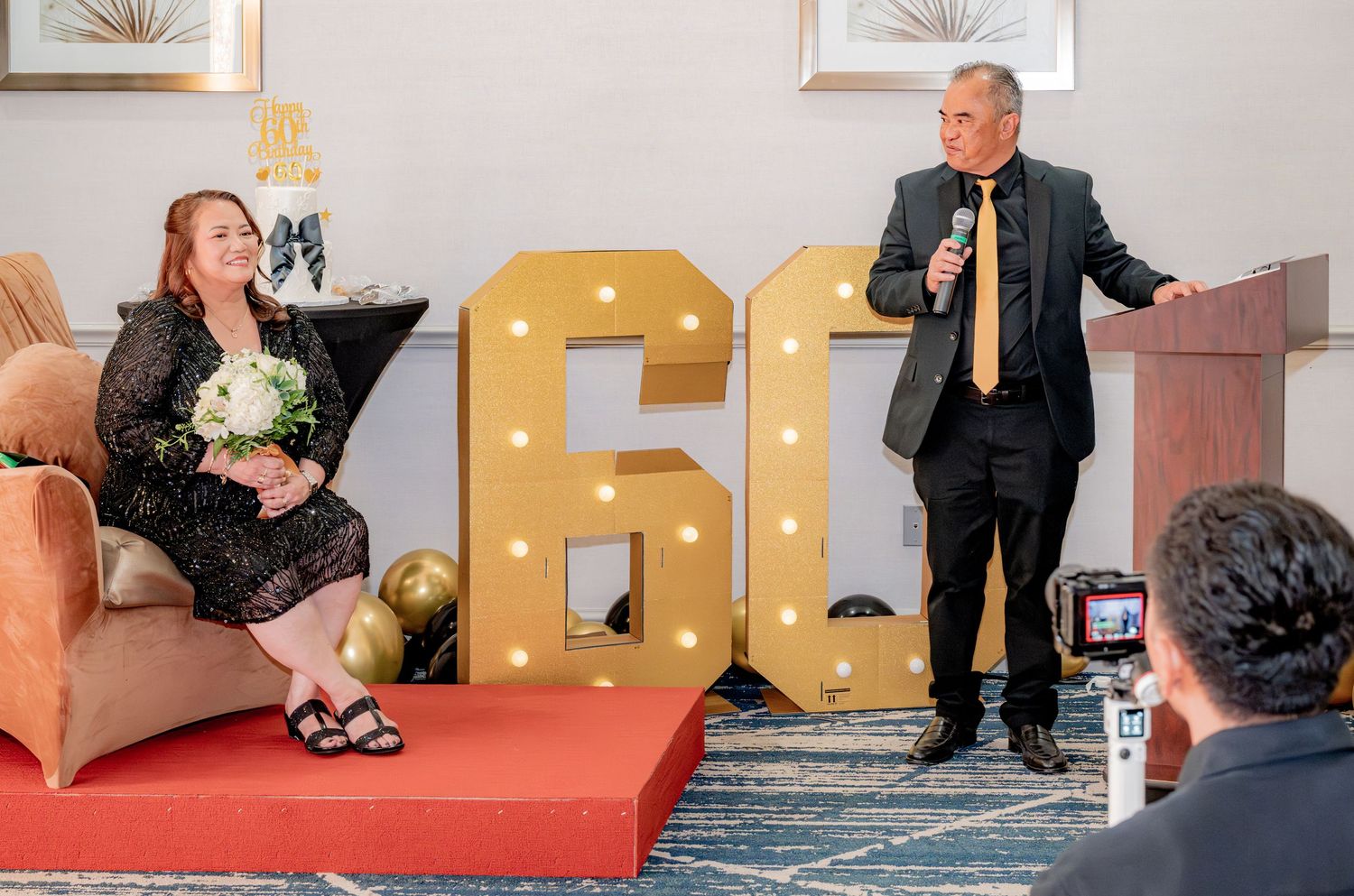 Man giving speech at podium beside illuminated 60 marquee sign at birthday party.