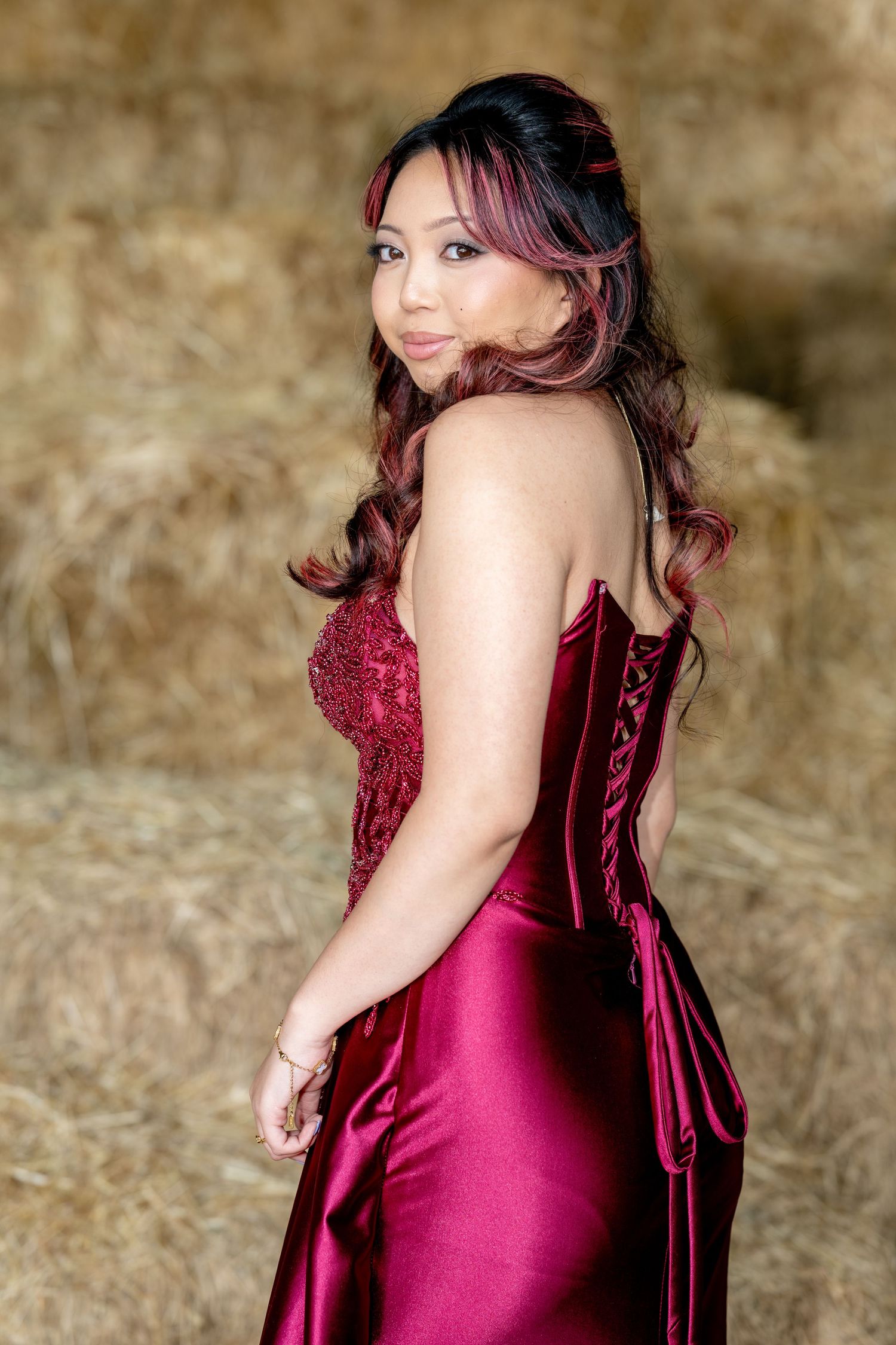 Woman in strapless red satin prom dress with corset back smiles over shoulder in rustic hay bale setting.