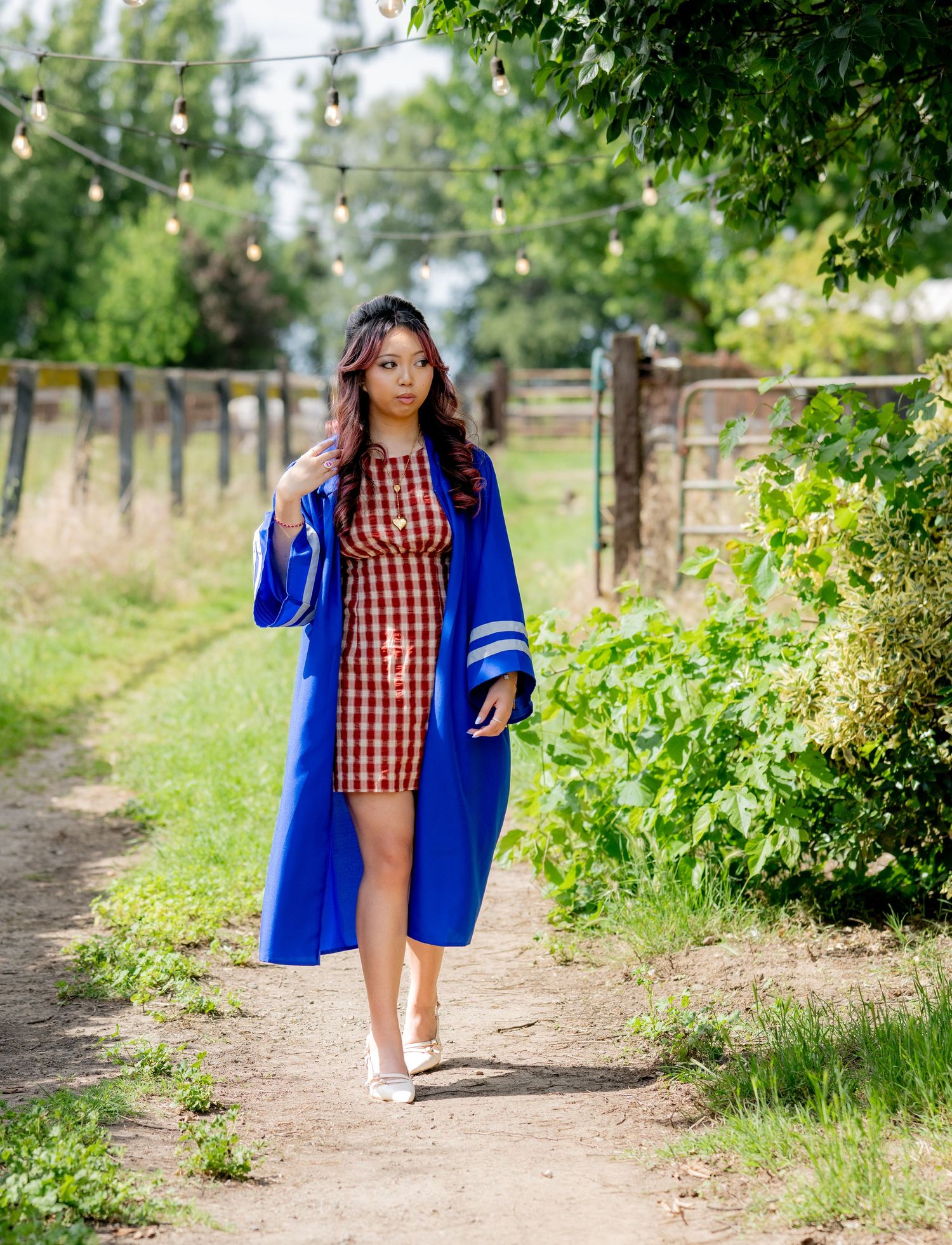 Young woman in blue graduation gown and red checkered dress walks sunlit outdoor path.