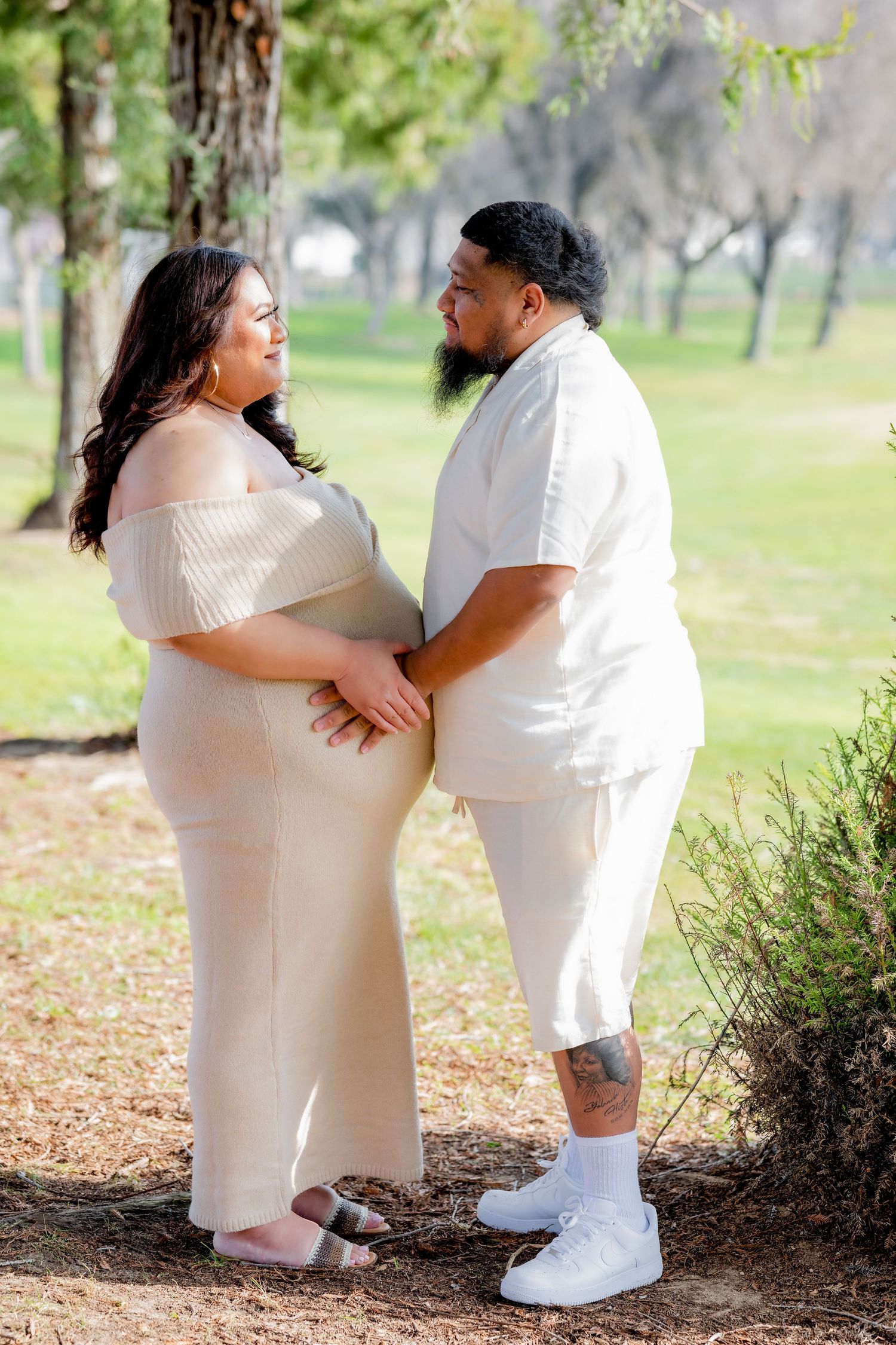 Expectant couple in matching cream outfits share a tender moment gazing at each other in a sunny park setting.