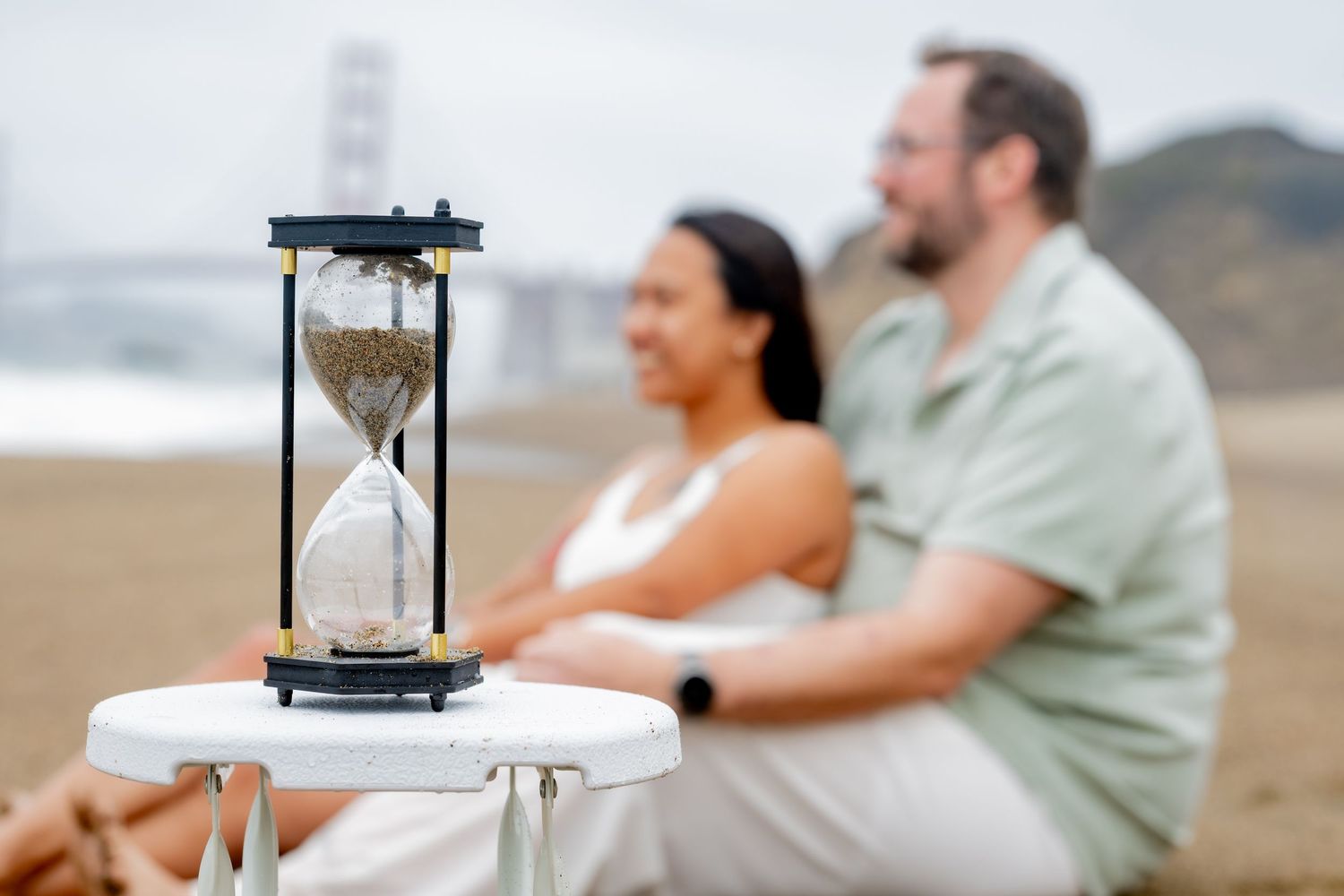 Hourglass on white stool in foreground with couple sitting together on Baker Beach behind it.