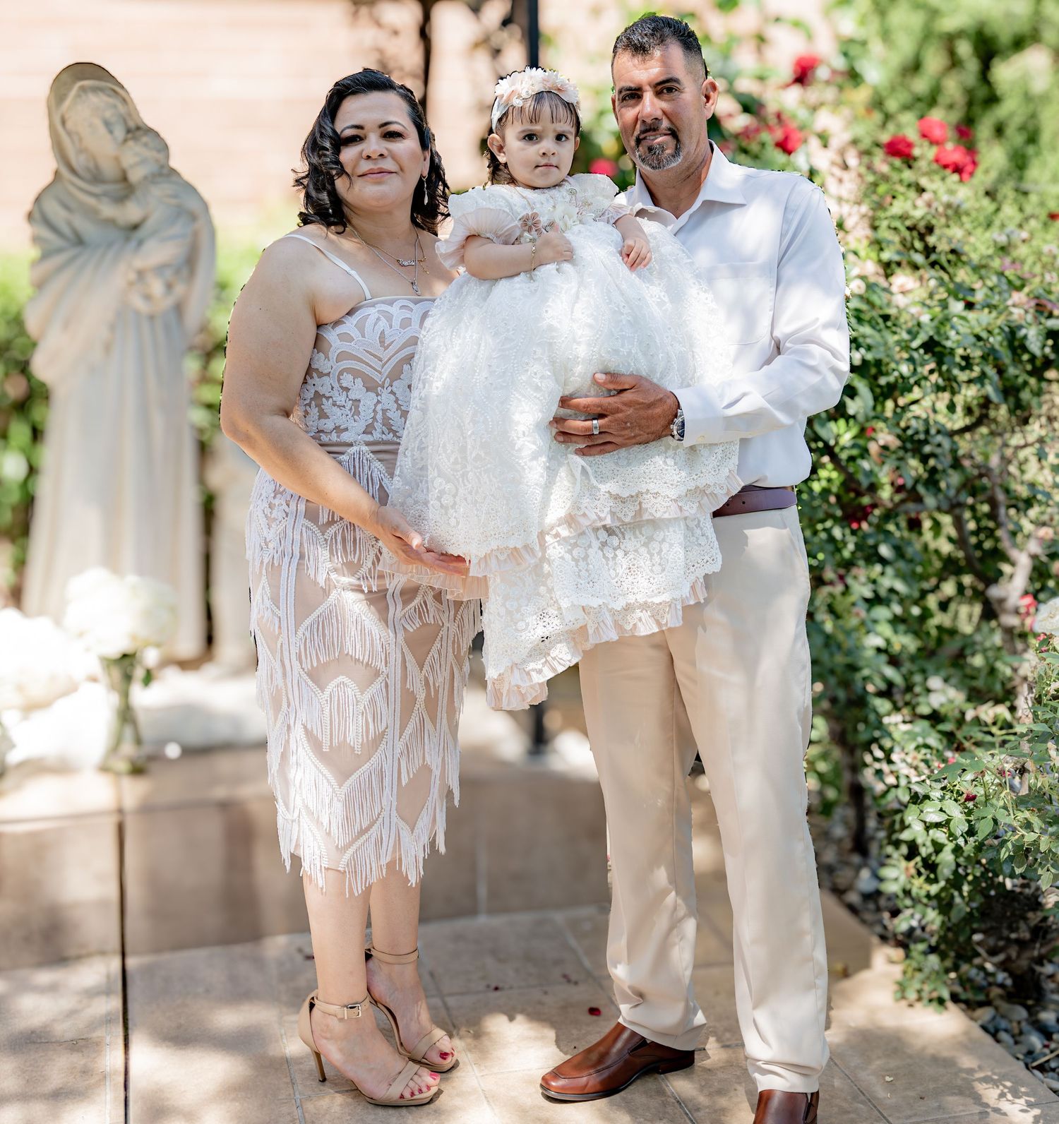 Couple in formal attire holds baby in white baptism gown near Virgin Mary statue in garden.