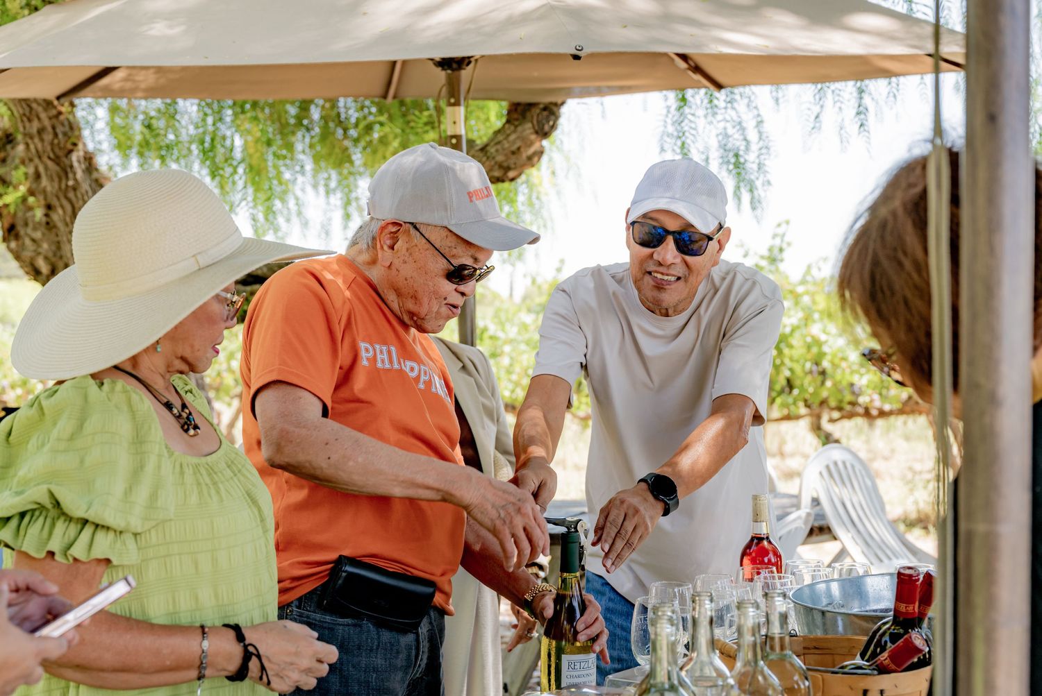 People enjoying an outdoor wine tasting event under a canopy, with a server pouring drinks on a sunny day.
