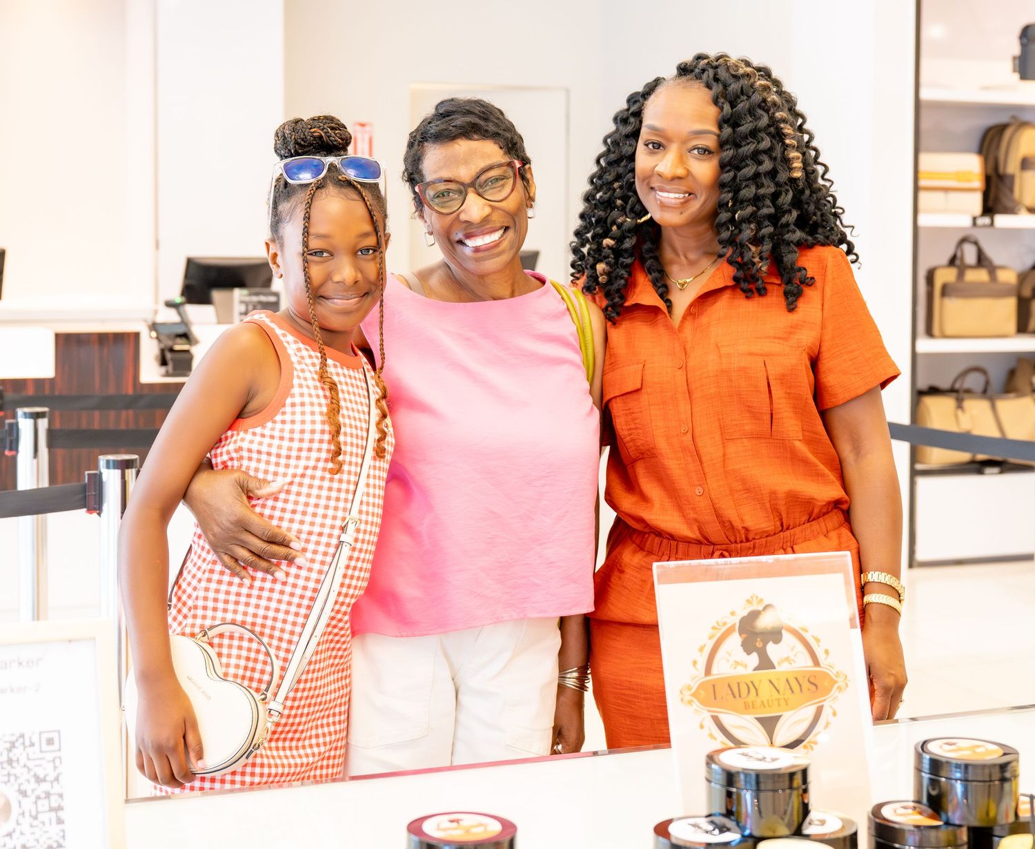 Three women smiling together in a bright retail store, wearing colorful outfits in orange and pink tones.