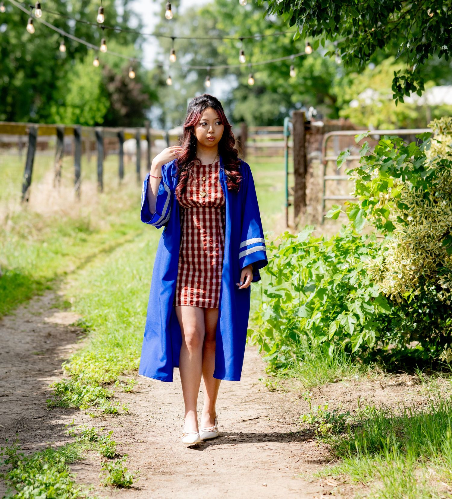 Young woman in blue graduation gown strolls sunny garden path with string lights above.