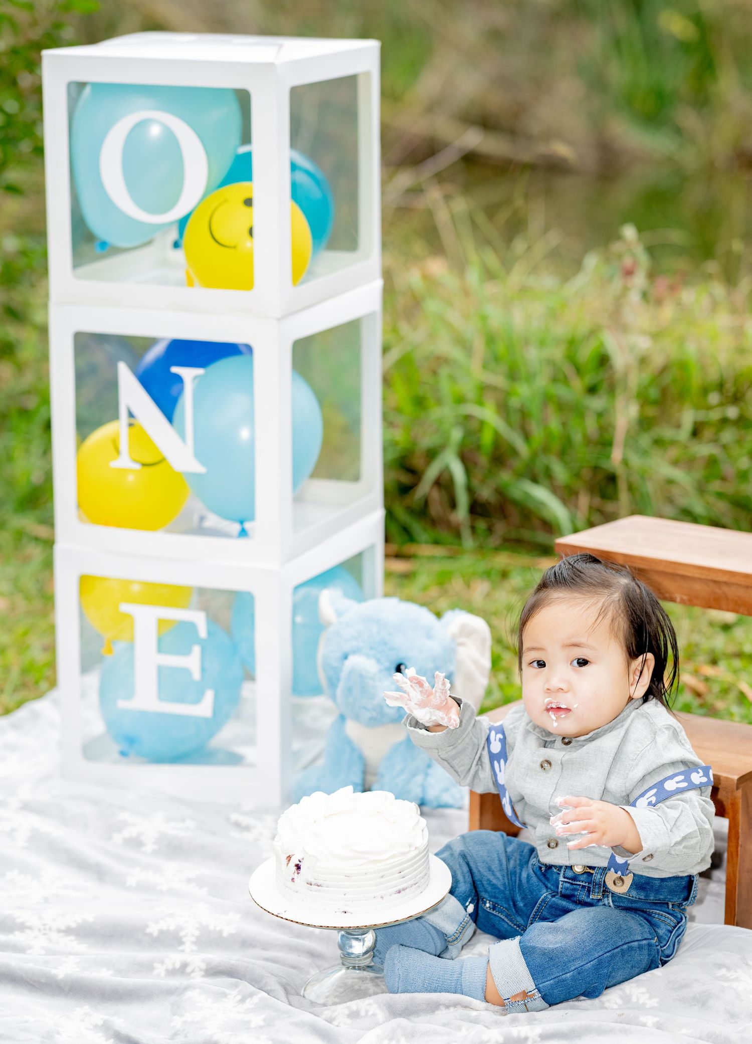Baby boy sitting with smash cake at first birthday outdoor photo shoot with ONE balloon boxes and blue teddy bear.