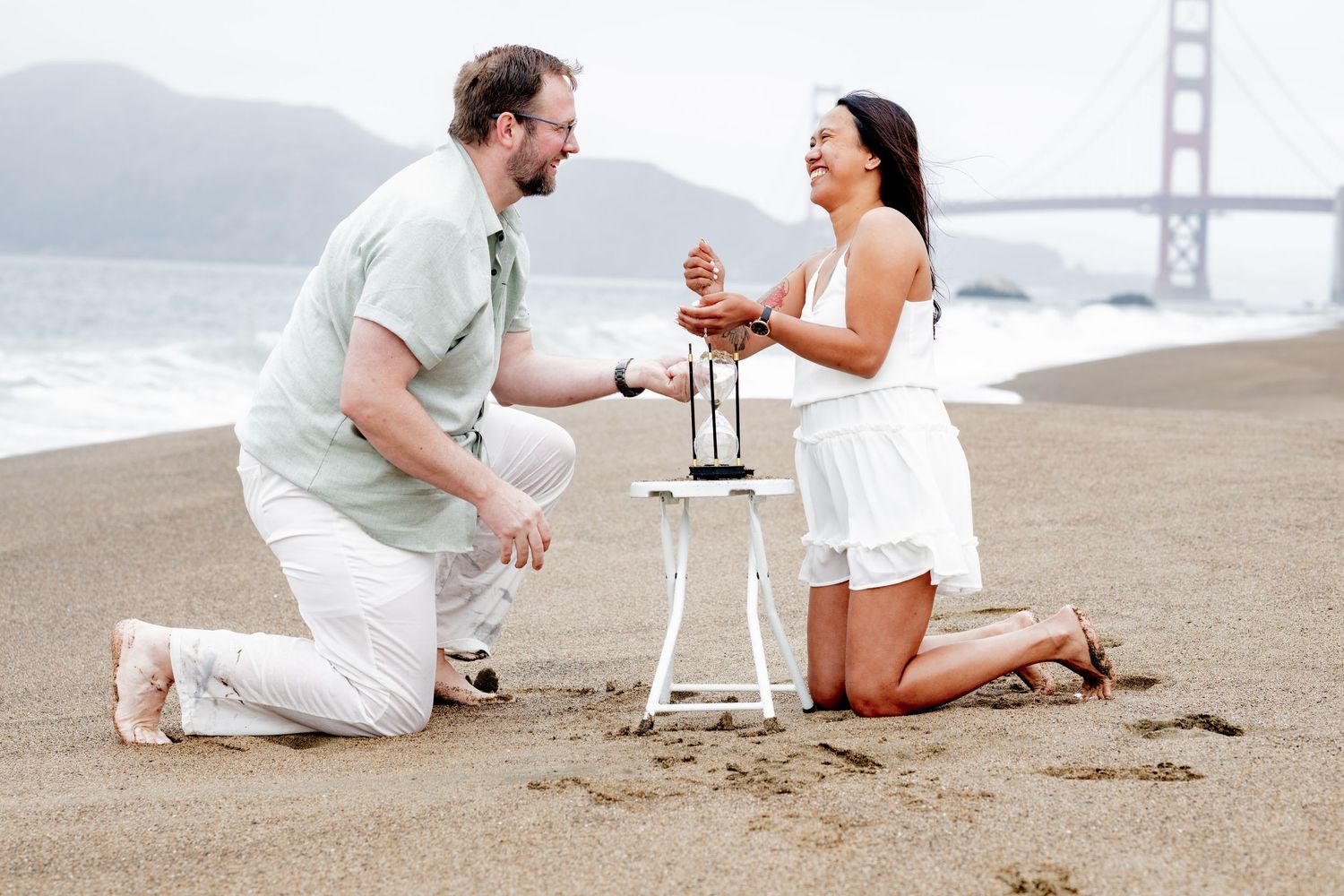 Couple kneeling on Baker Beach with Golden Gate Bridge, sharing a romantic moment over a lantern.