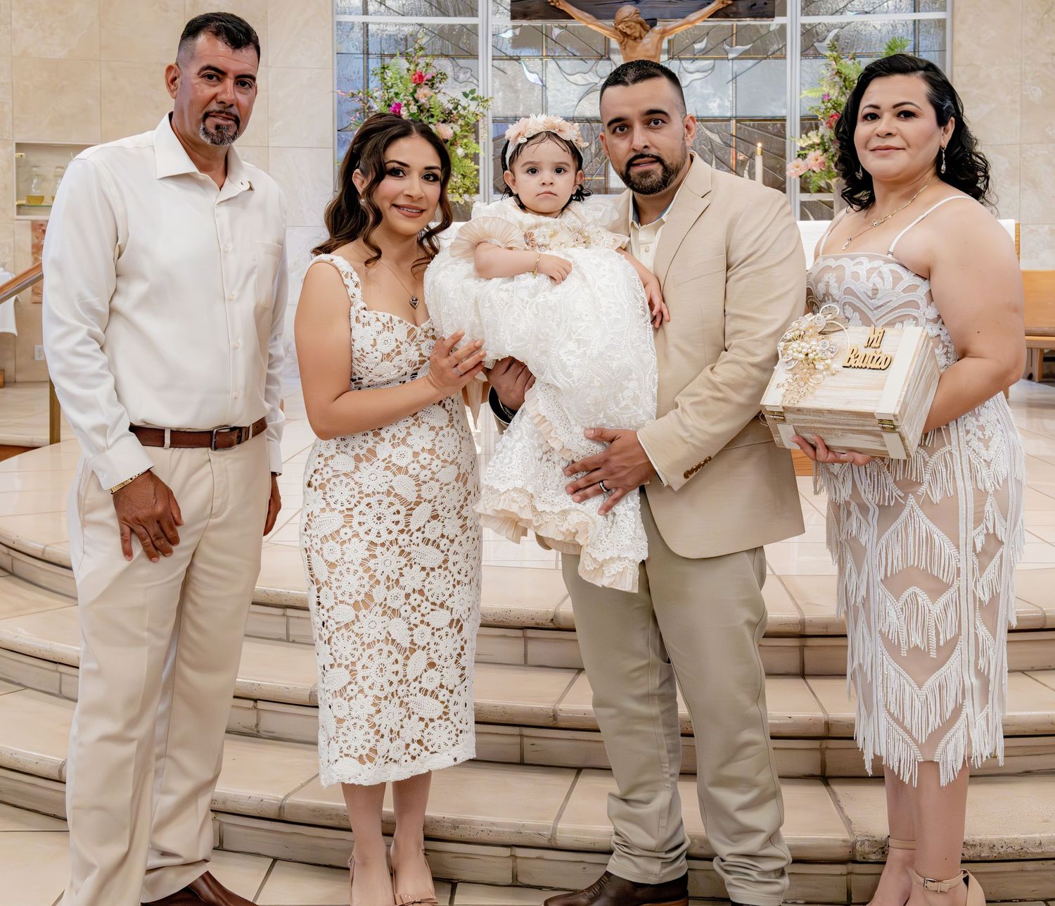 Family of five poses with baptized baby in white gown at church altar steps.