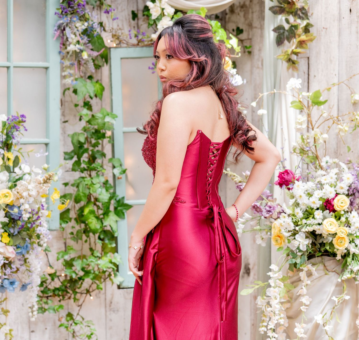 Elegant back view of woman in deep red satin gown with lace-up corset back in floral decorated venue.