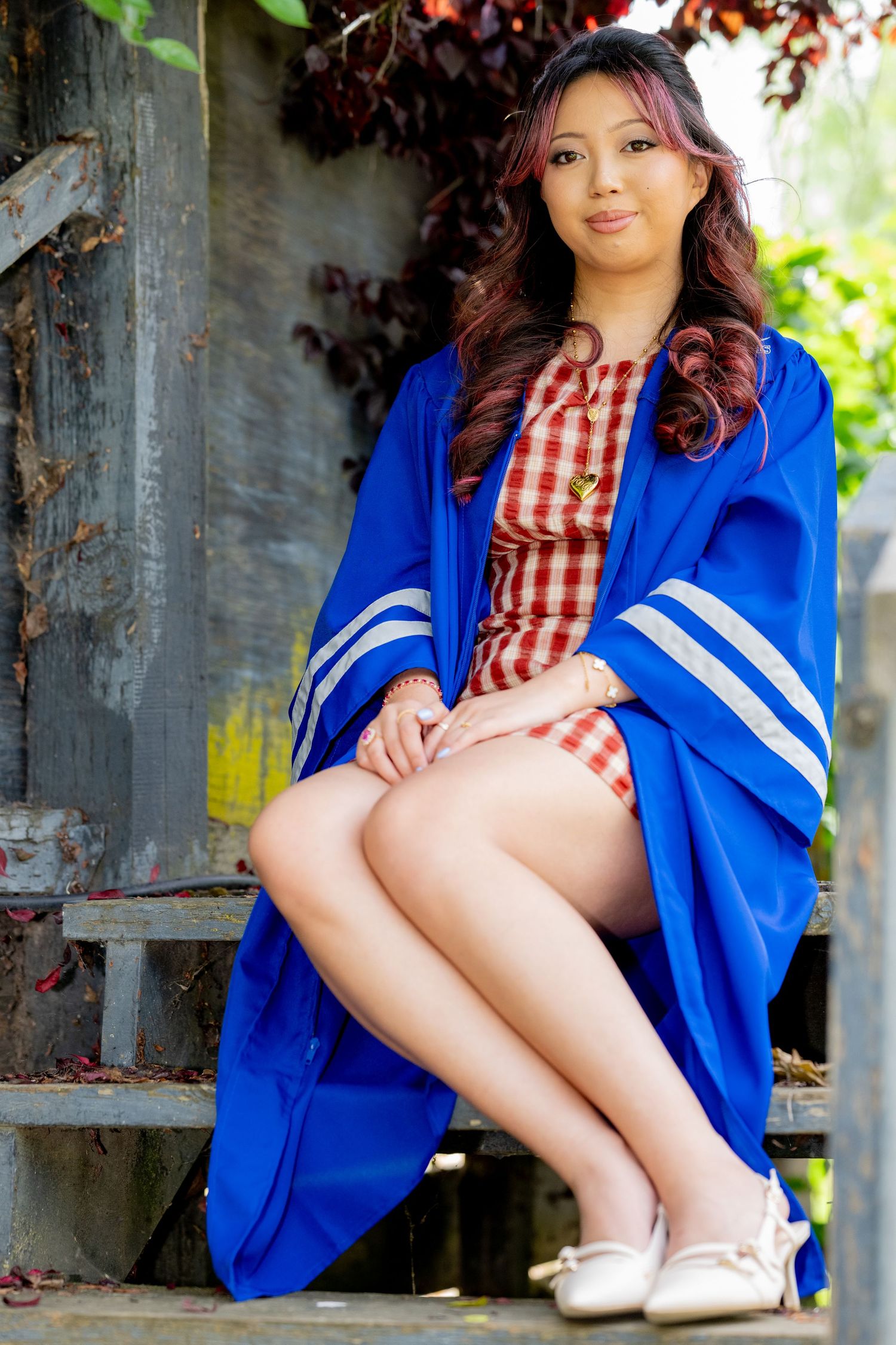 Smiling graduate in blue gown and red gingham dress sits on steps in lush garden setting.