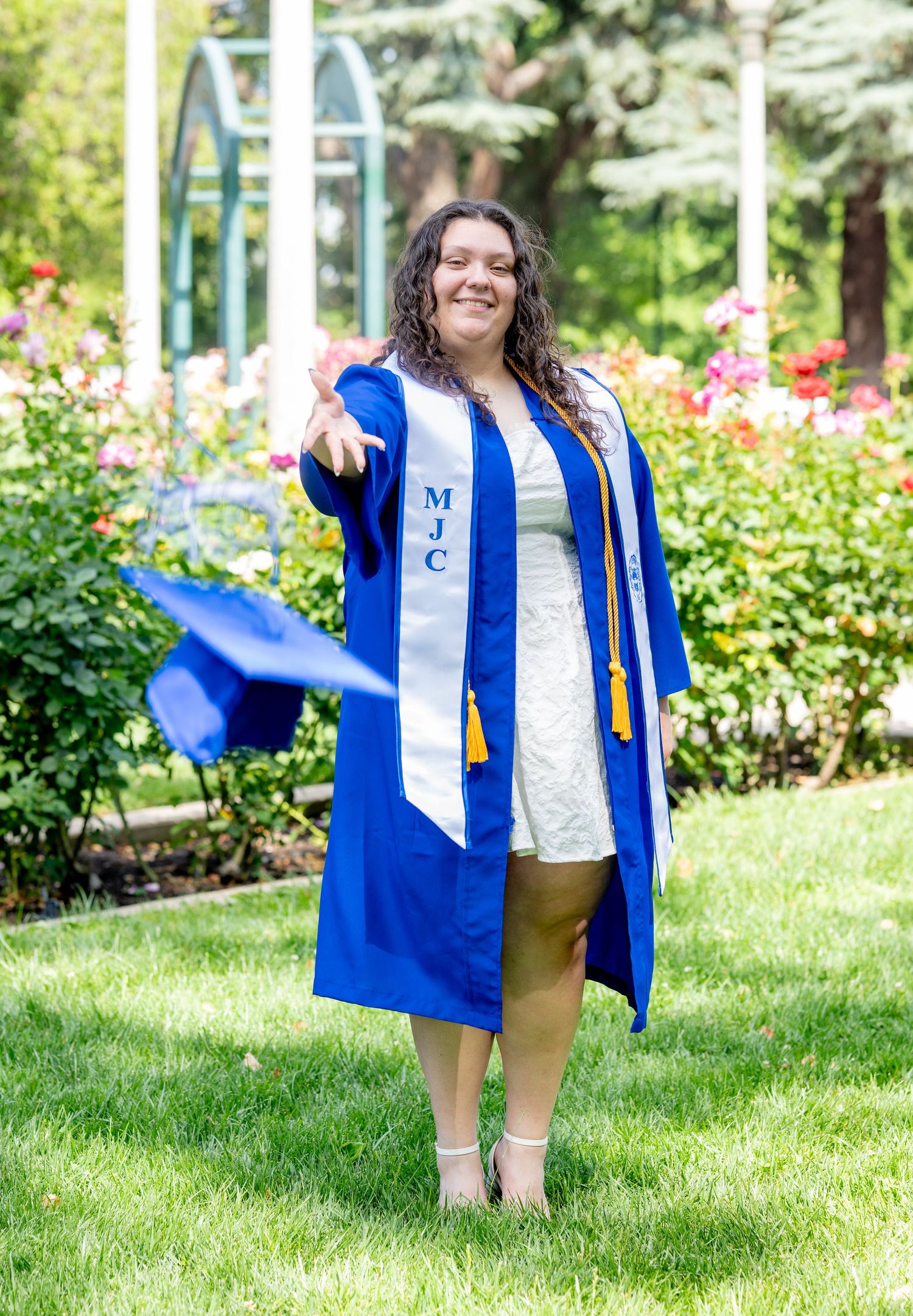 Woman in blue MJC graduation gown and stole tosses cap in the air outdoors.
