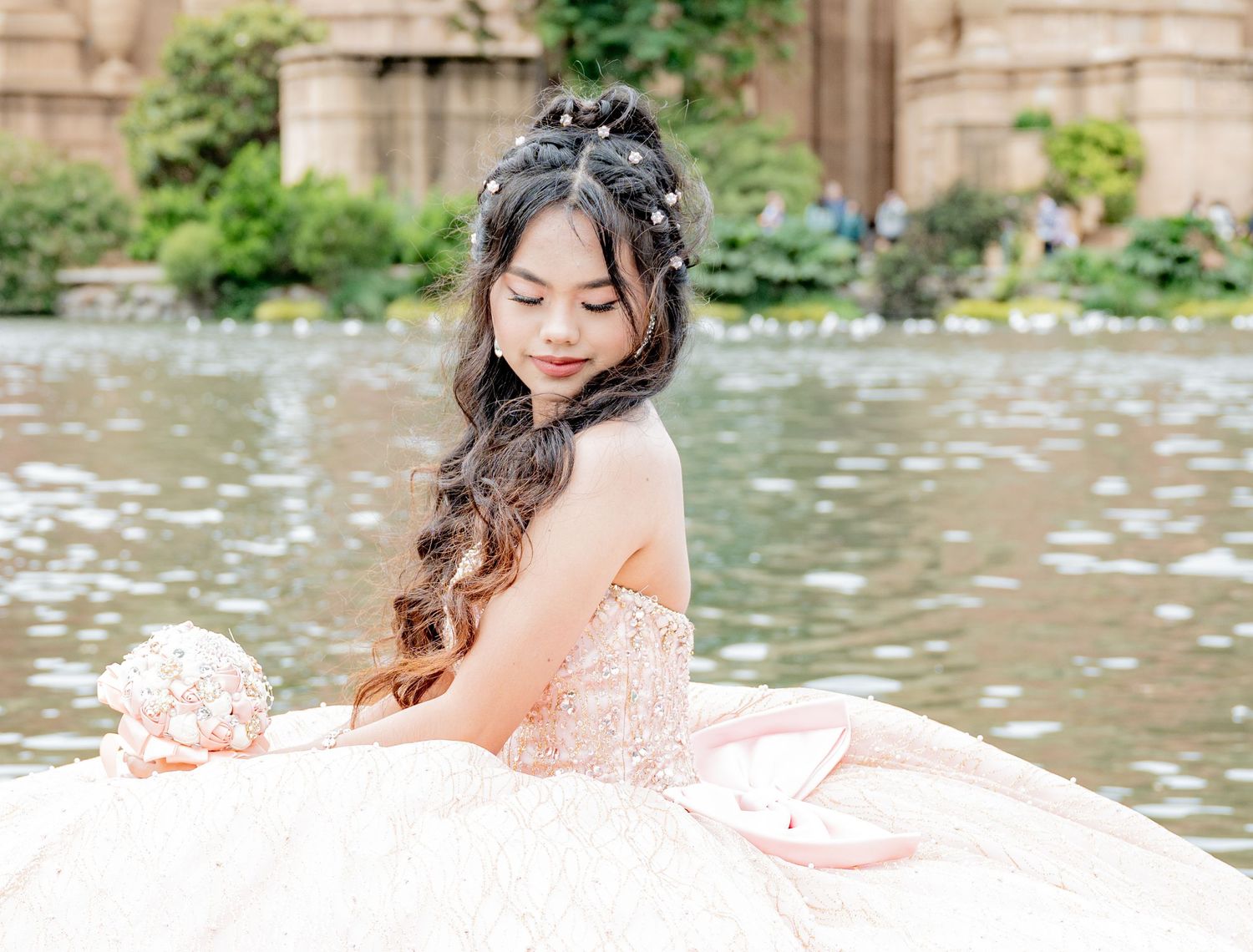 Young girl in voluminous blush ball gown sitting by water holding decorative bouquet.