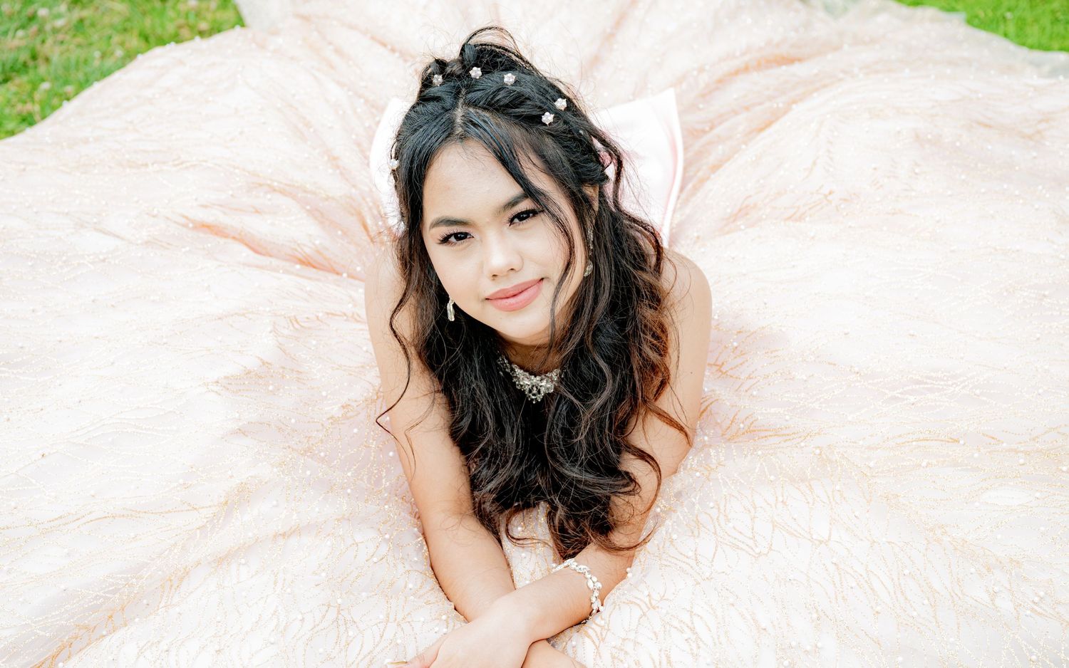 Young girl in elegant blush ball gown lying on grass, wearing pearl crown, smiling.