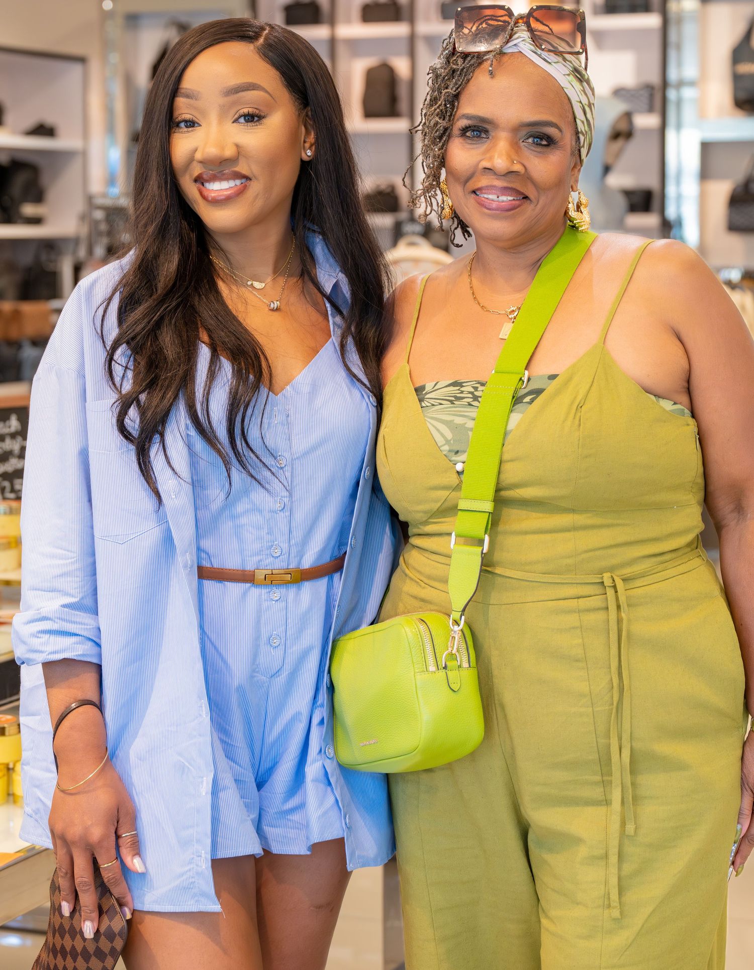 Two stylish women posing together in a store, one in a blue dress and one in a green outfit with matching bag.