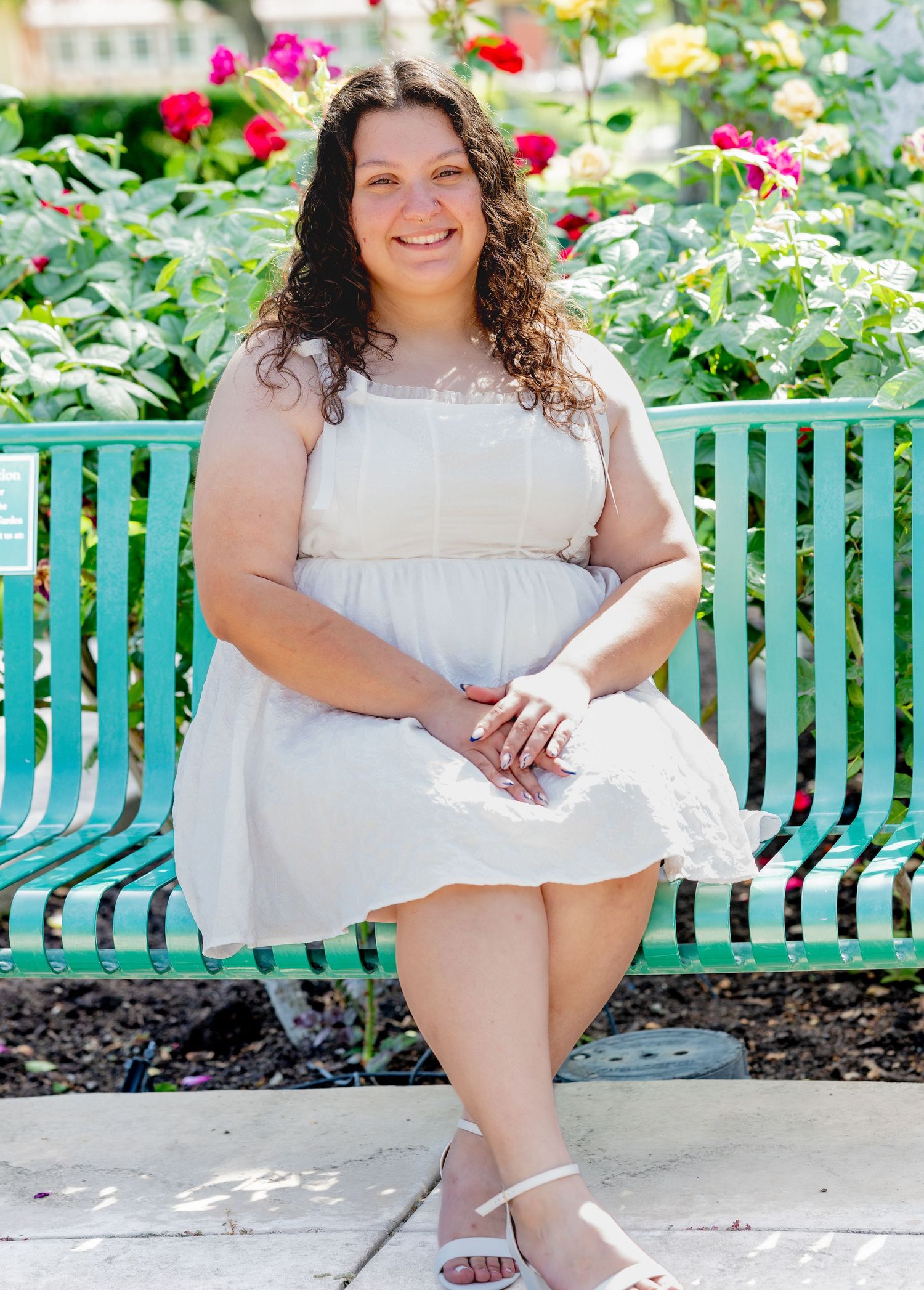Young woman in white dress sitting on green bench surrounded by colorful rose bushes.