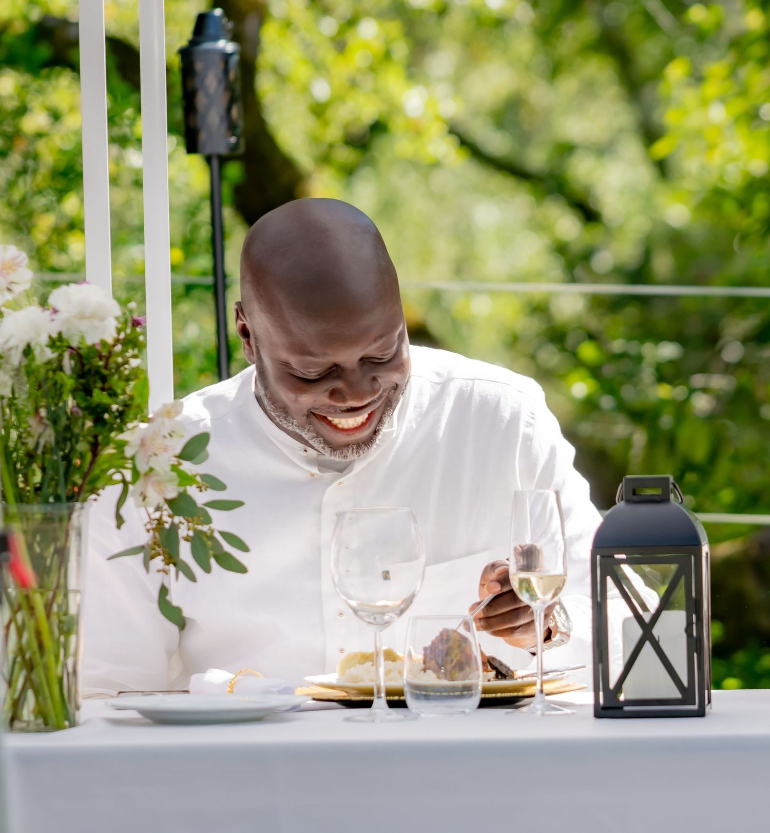 Smiling man in white shirt enjoying outdoor dining at elegant table with lantern and flowers.