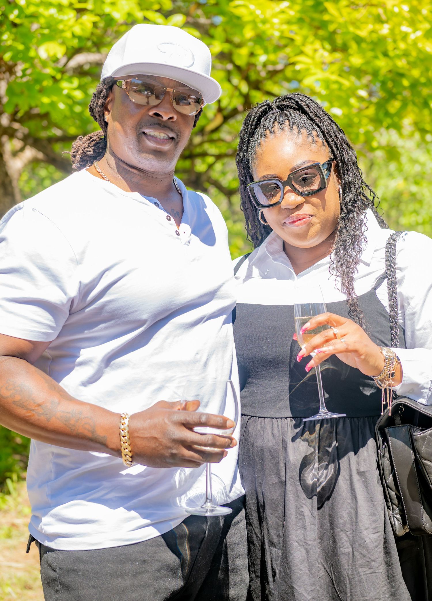 Couple posing outdoors at a garden party, man in white cap holding champagne glass with woman in sunglasses.