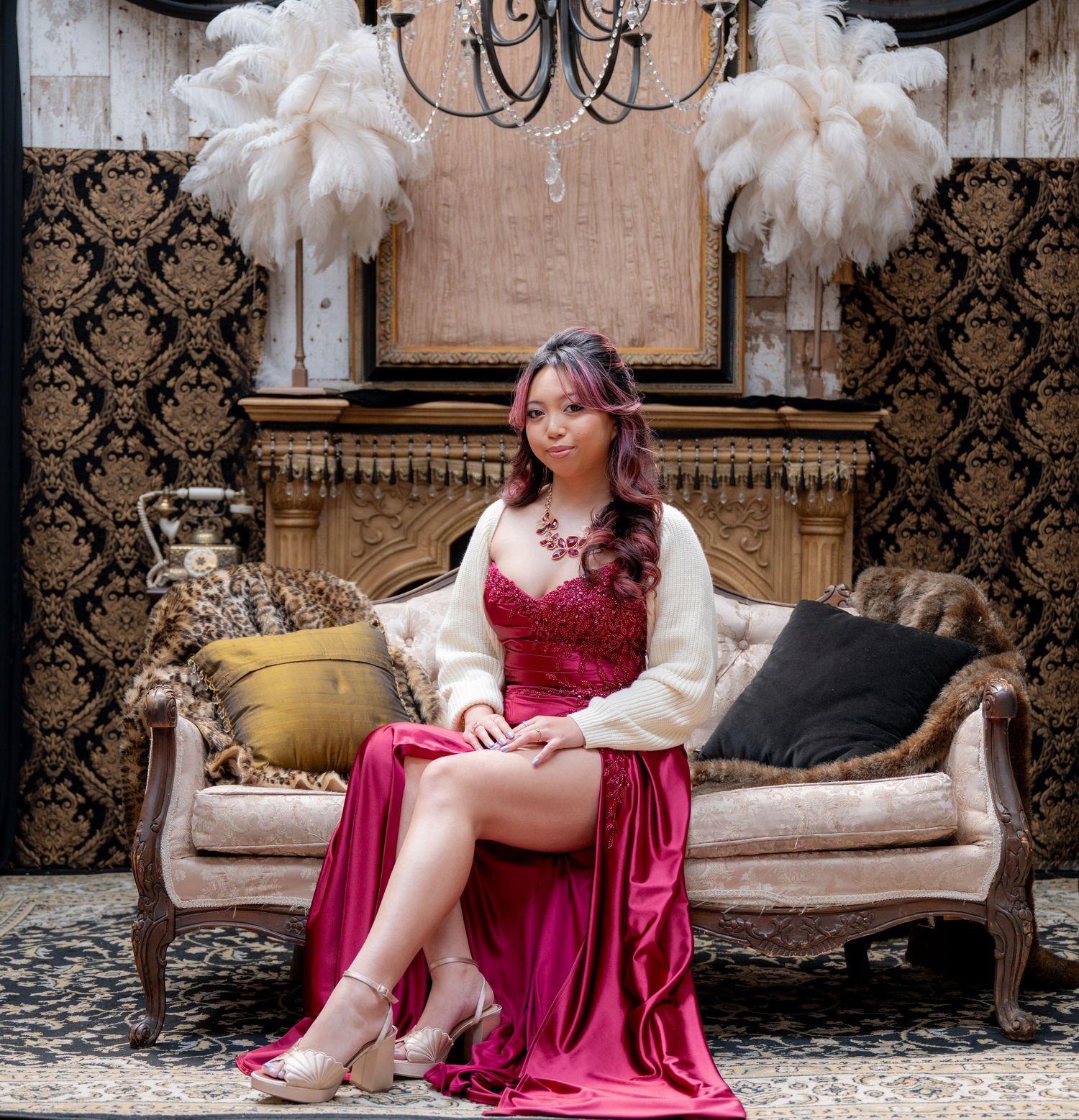 Young woman in red satin gown and white cardigan seated on vintage sofa with feather decor.