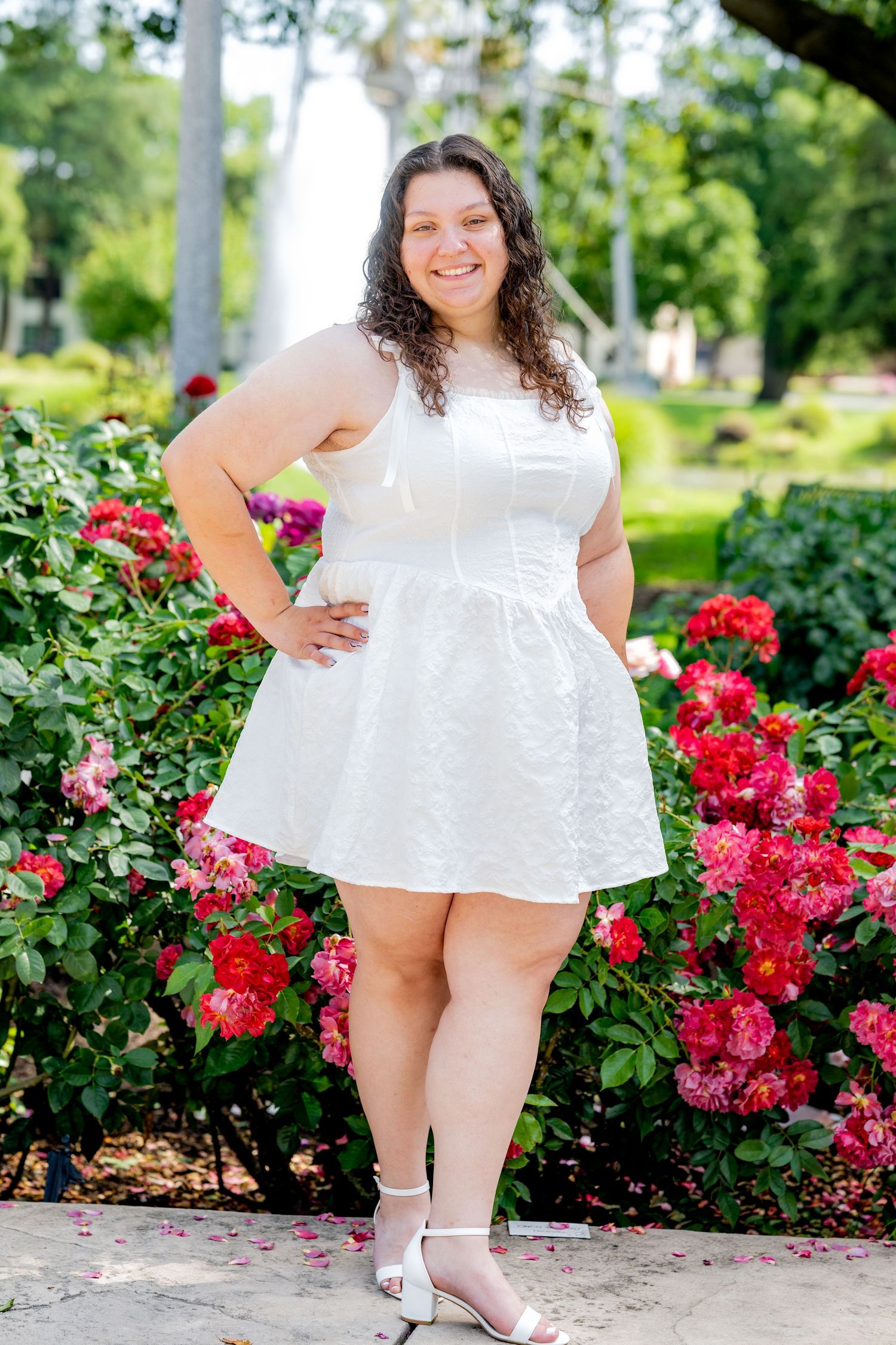 Young woman in white dress smiling confidently in front of vibrant red rose bushes.