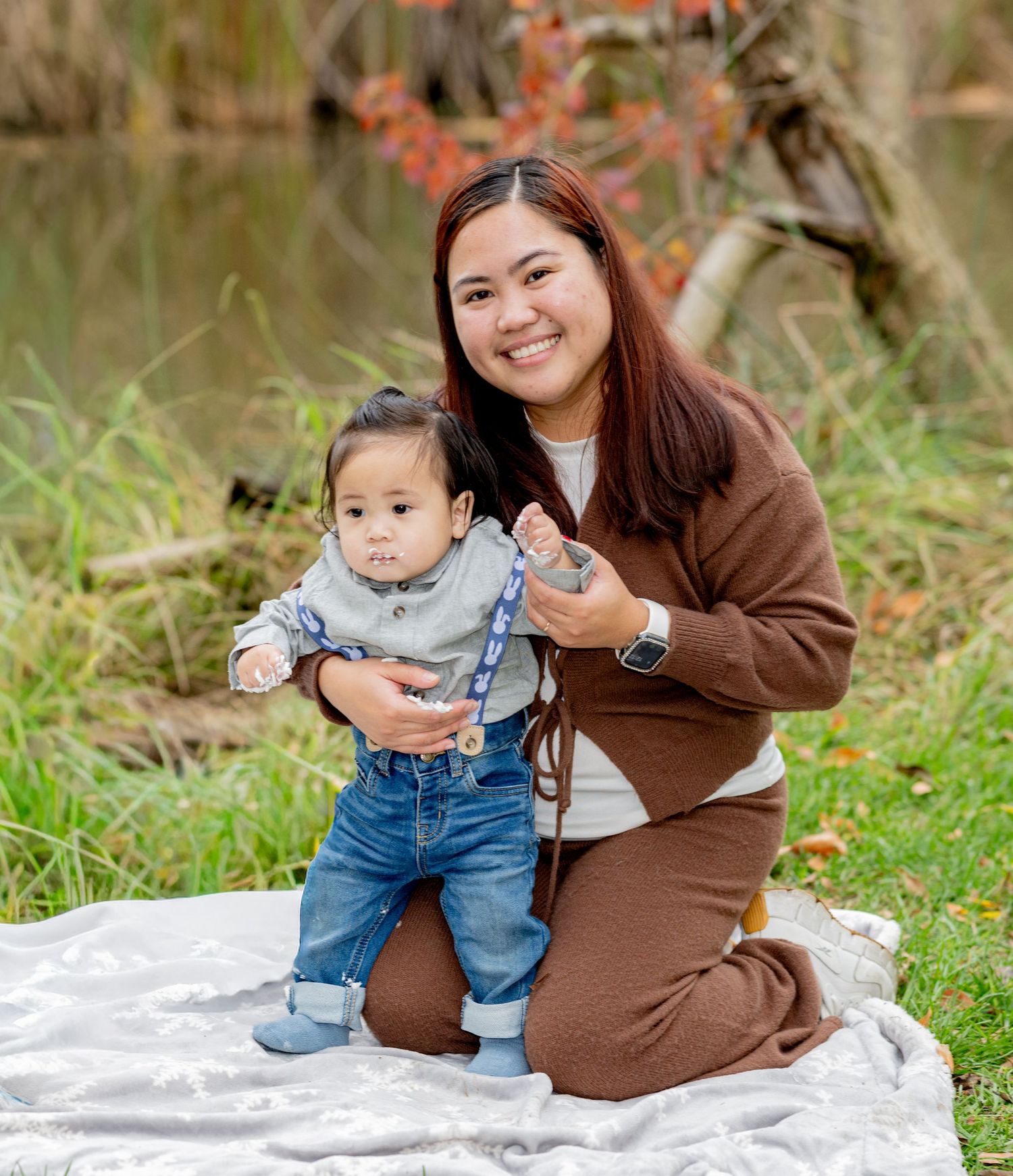 Mother kneeling behind standing baby boy on white blanket during outdoor birthday photoshoot in park.