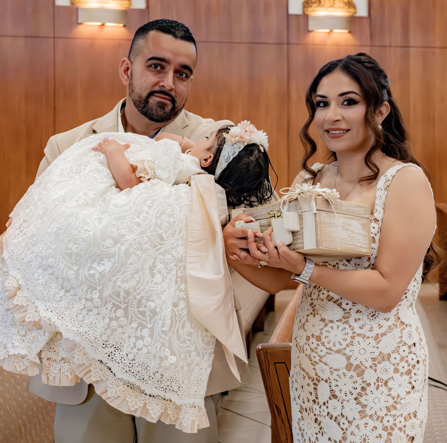 Proud parents pose with baby in ornate white lace baptism gown and decorative gift box during christening ceremony.
