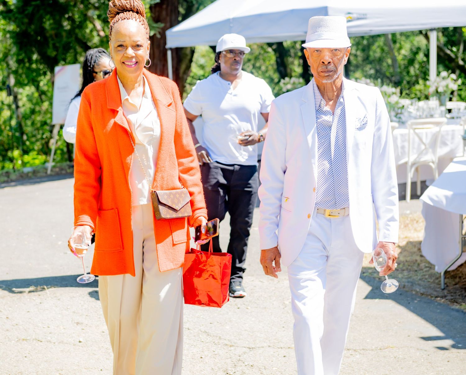 Elegant couple walking outdoors, woman in orange blazer and man in white suit with hat at a garden event.