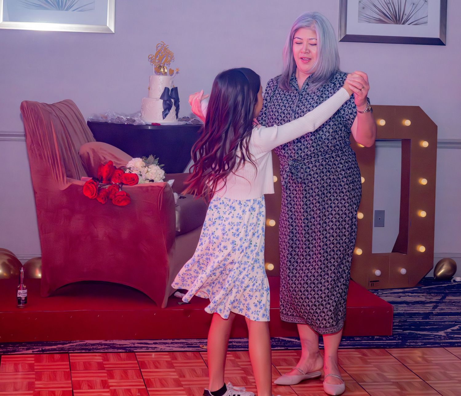 Woman in patterned dress dancing with young girl near wedding cake and marquee lights.
