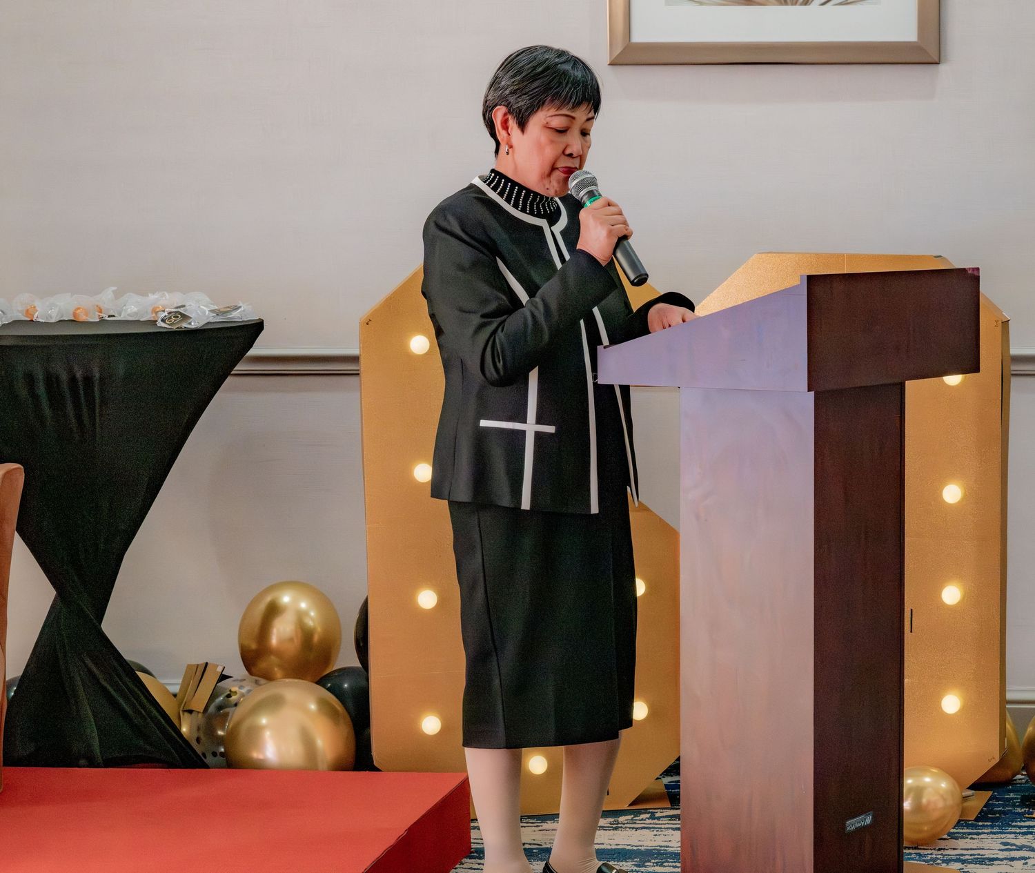 Woman in black suit speaking at a podium with gold balloon decorations at a formal birthday event.