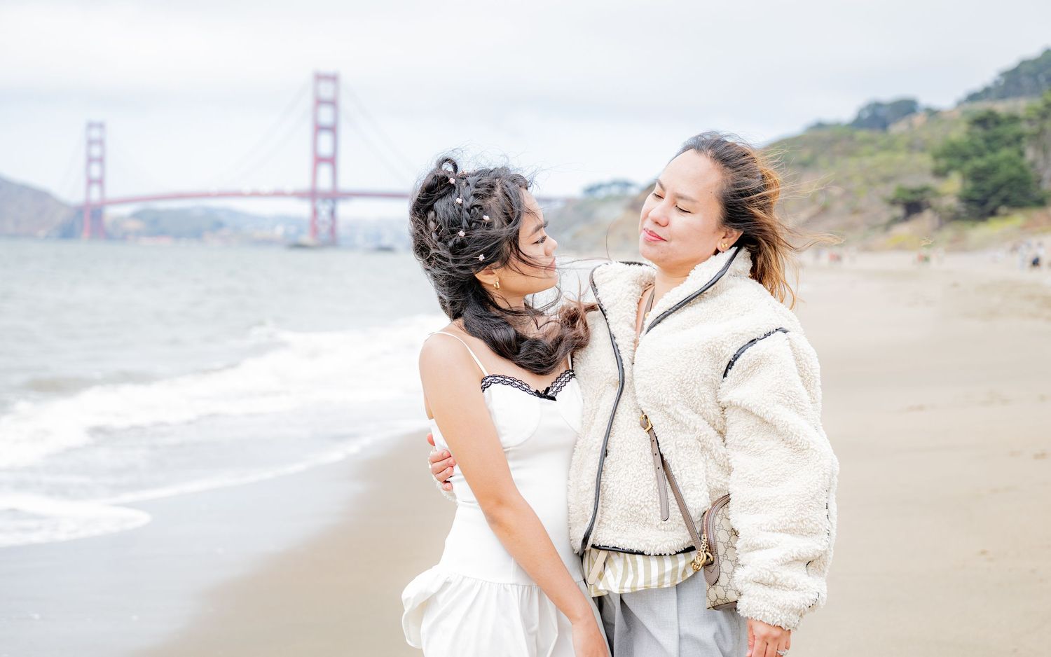 Two young women laugh together on a beach with the Golden Gate Bridge visible behind them.