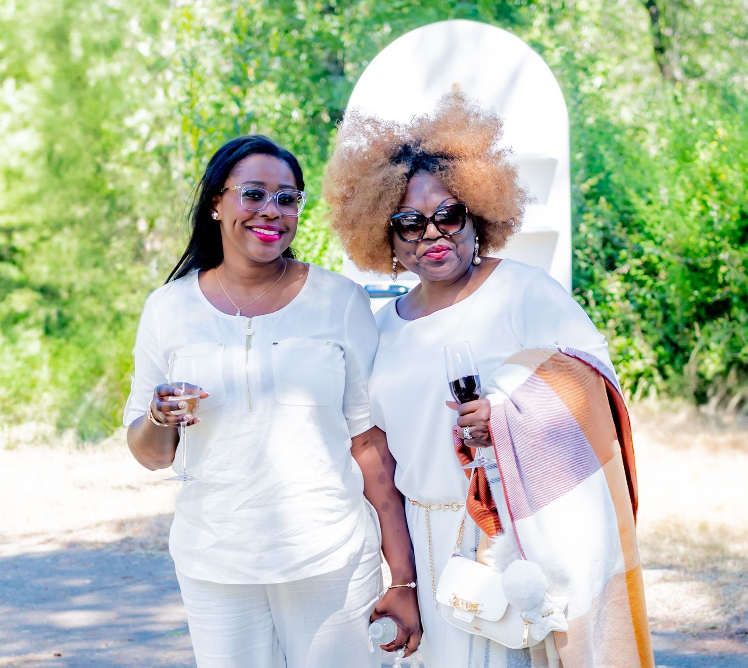 Two women in white outfits smiling outdoors at a sunny garden event.