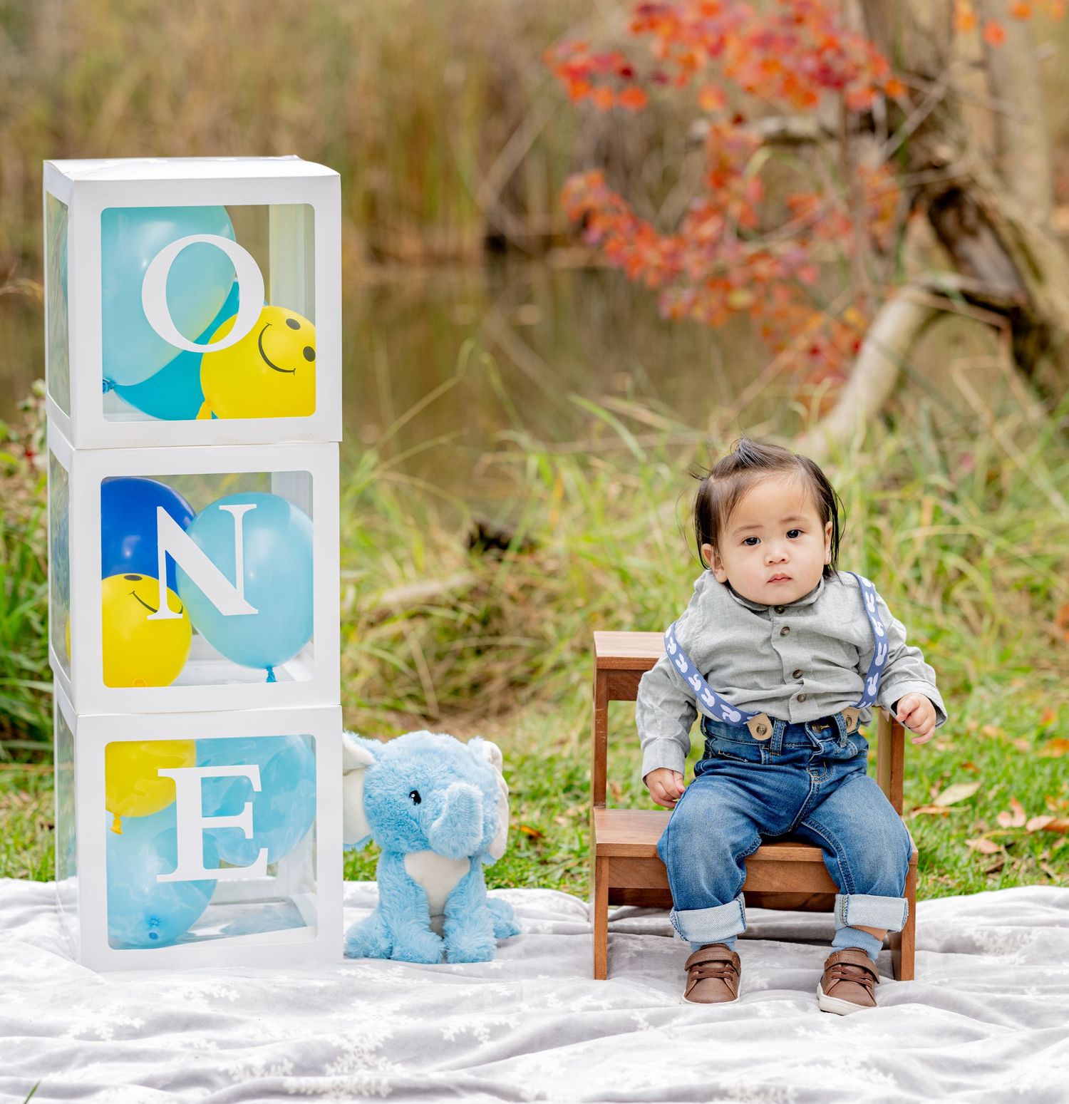 One-year-old boy sits beside stacked ONE balloon boxes with blue elephant plush toy outdoors.