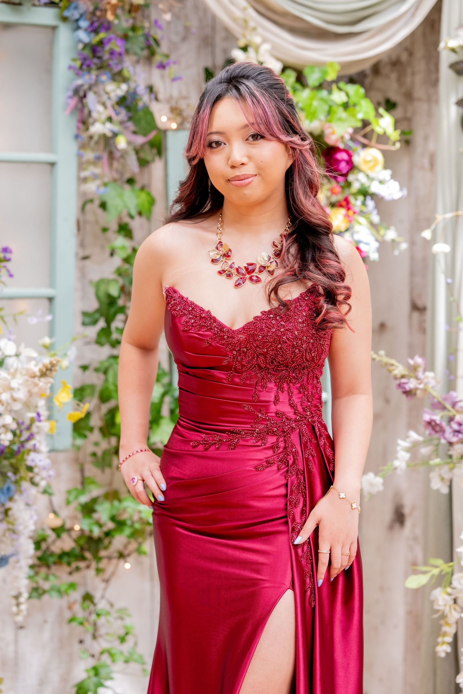 Young woman in elegant red lace strapless gown with floral necklace posing before a floral backdrop.