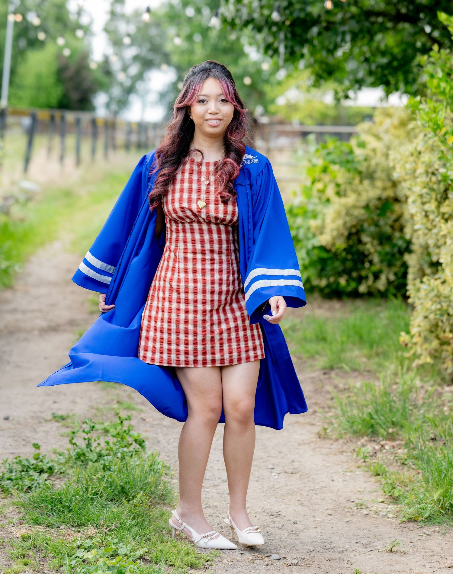 Smiling graduate in royal blue gown and red plaid dress poses on a sunny outdoor garden path.