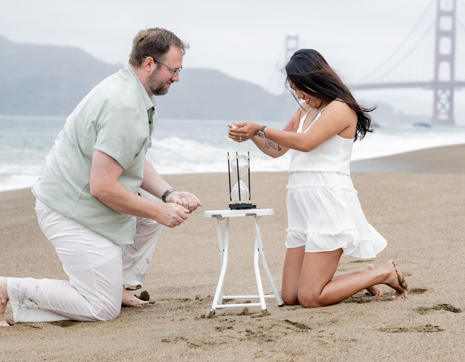 Couple kneeling on Baker Beach examining a small decorative table with Golden Gate Bridge behind.