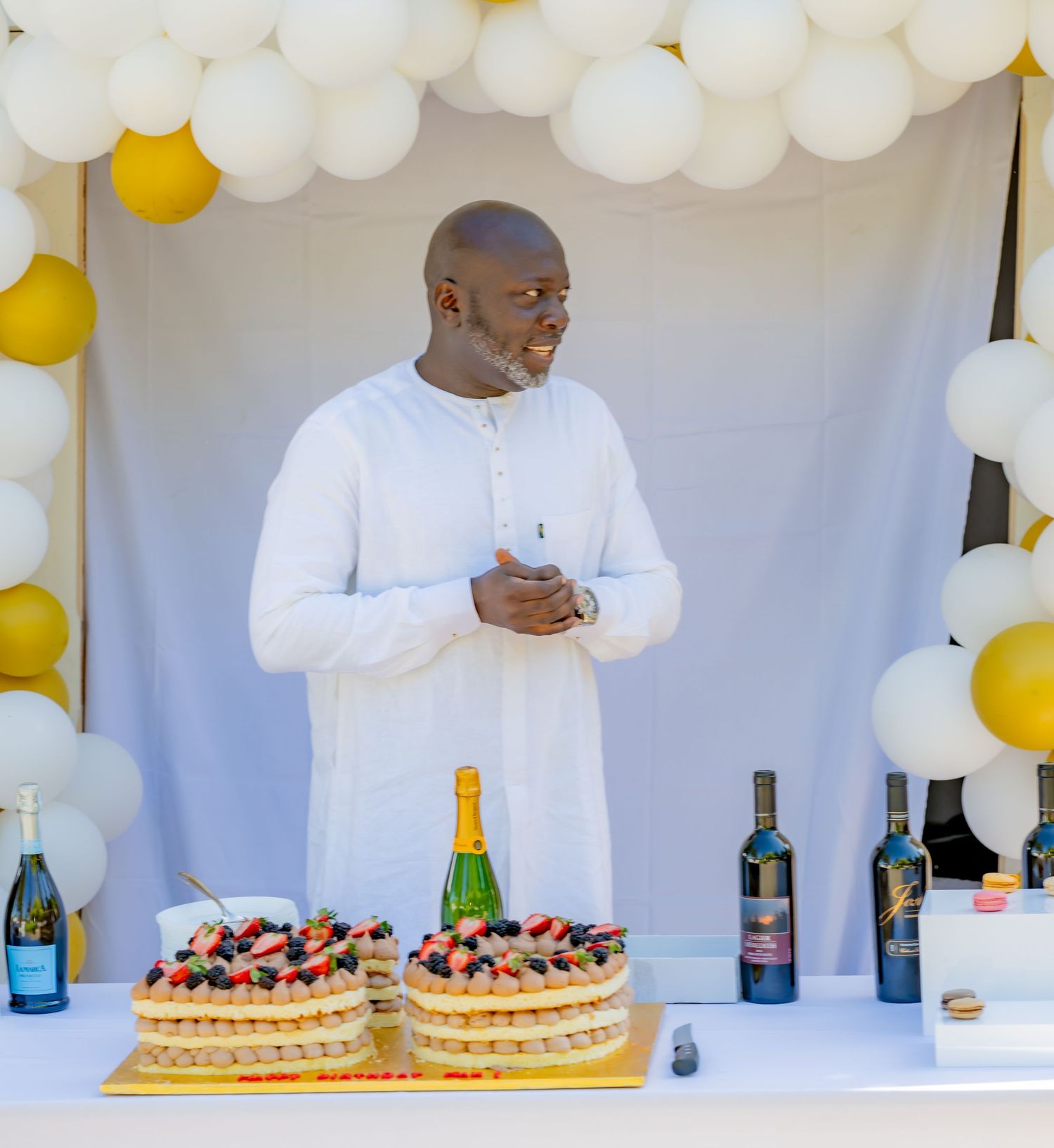 Man in white outfit standing behind a dessert table with cakes, wine, and balloon decorations.