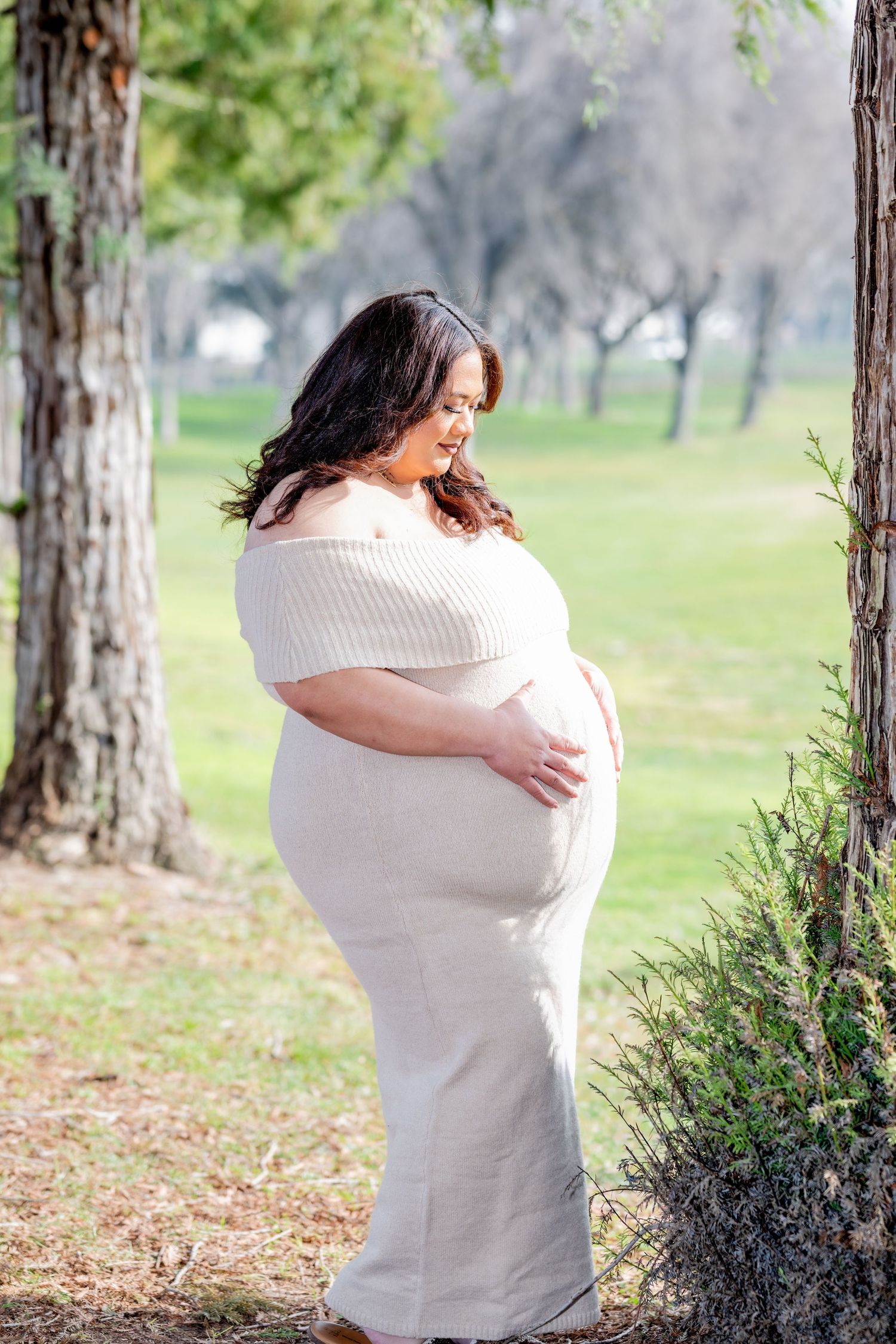 Pregnant woman in cream off-shoulder dress gently holds her baby bump while standing near trees in a park.