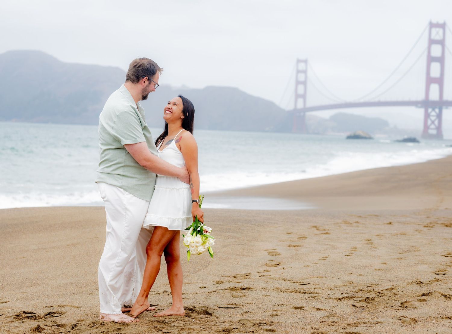 Happy couple smiling at each other on Baker Beach with Golden Gate Bridge in the background.