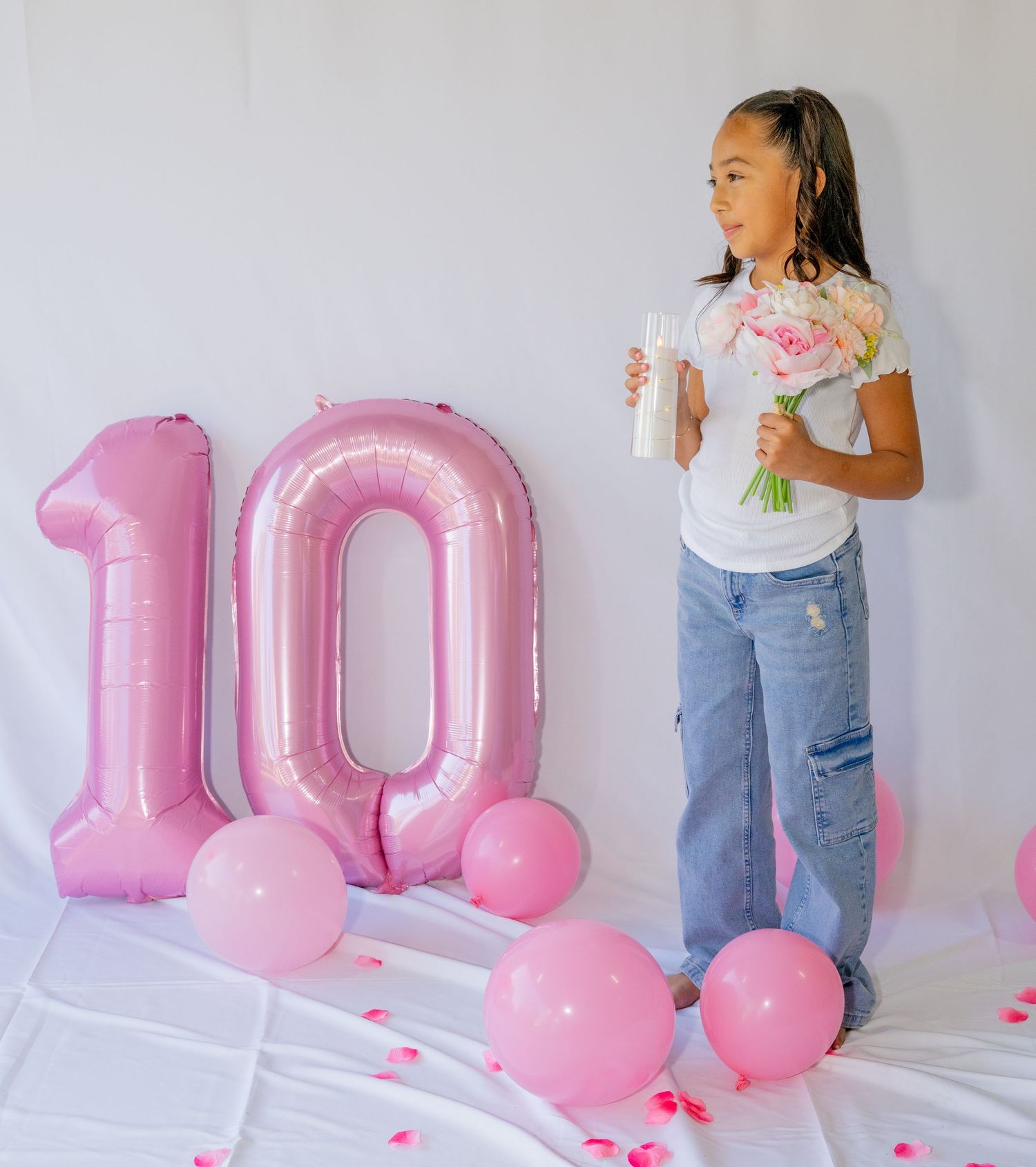 Girl stands beside pink number 10 balloon holding bouquet and candles on white backdrop.